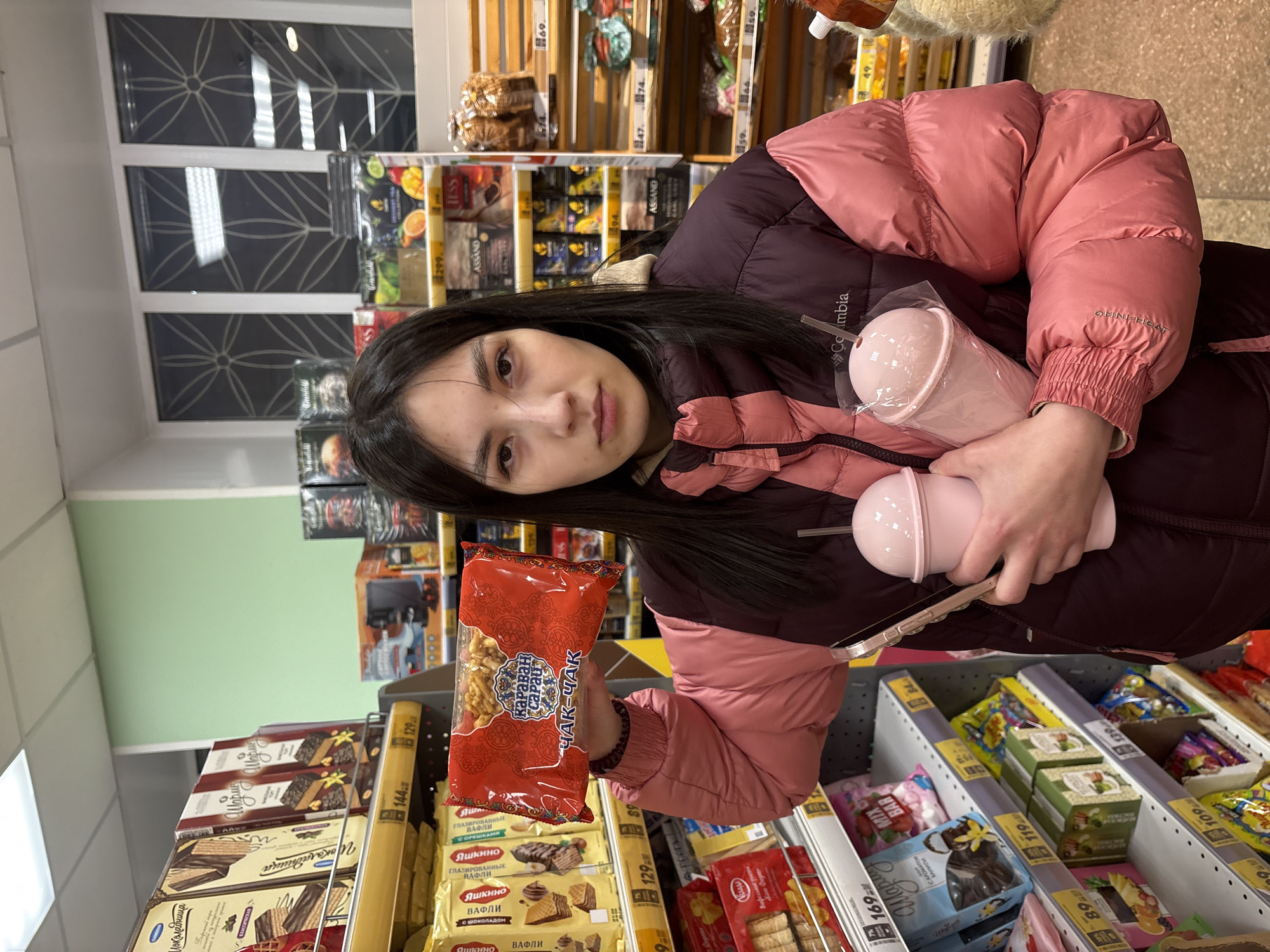 A young woman is standing in a grocery store in Chaikovsky, Russia, holding two pink tumblers and a package of "Chak-Chak" (a Tatar sweet pastry). She's wearing a maroon and pink Columbia winter jacket. She is in an aisle with chocolate and candy products. The shelves are stocked with items labeled in Russian, including "Yashkino" waffles and "Assand" tea. The prices are marked in rubles. There is a window in the background.