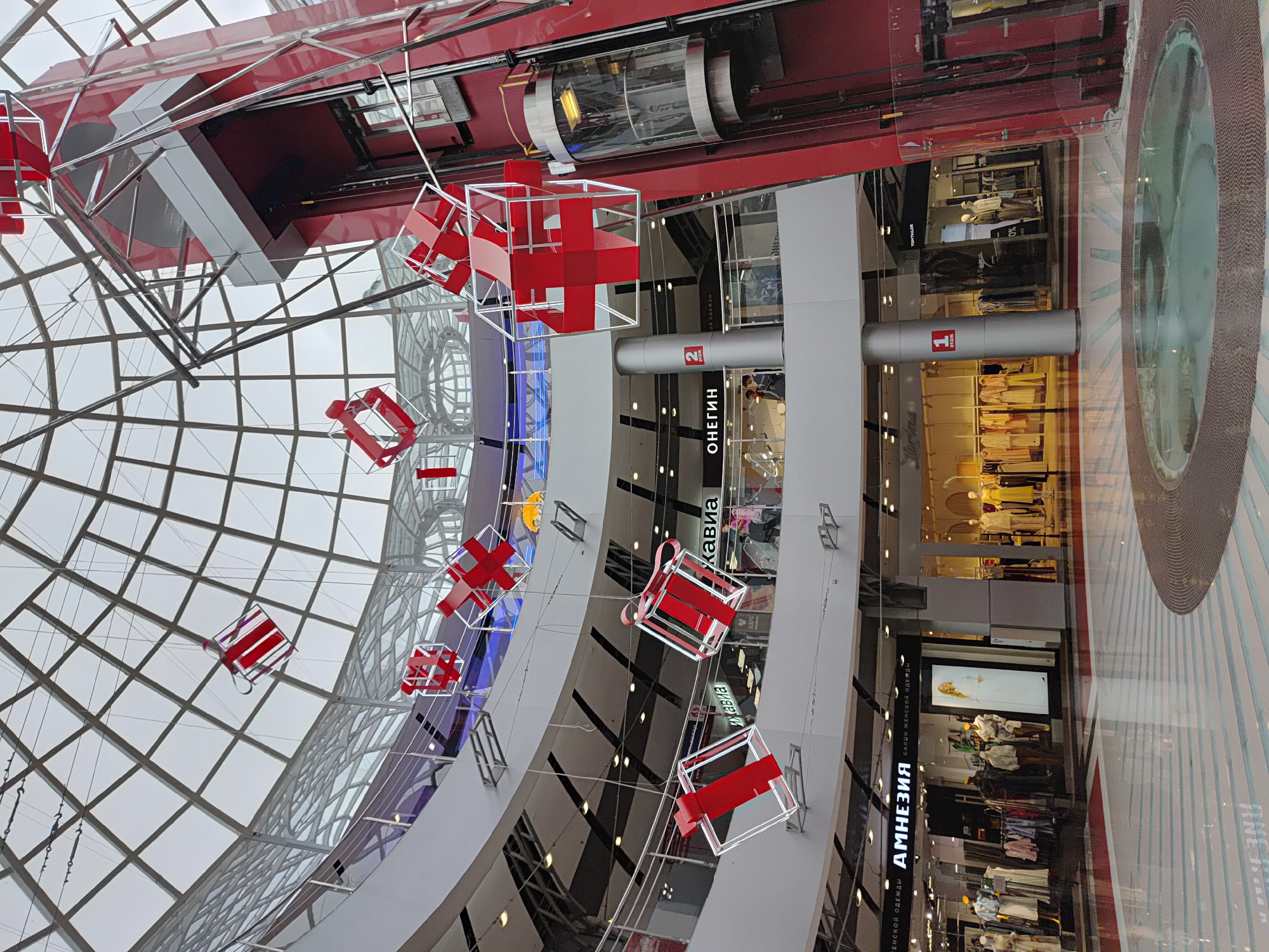 The image shows the interior of the "Talisman" shopping mall in Izhevsk, Russia. The architecture features a modern design with a glass ceiling, multi-level floors, and a cylindrical glass elevator. Red and white decorative structures hang from the ceiling, creating a festive atmosphere. Retail stores, identified by signs in Russian, line the floors. Clothing displays and mannequins are visible behind the store windows. Two silver pillars with the numbers "1" and "2" are visible at the top of stores respectively. The image is taken from a low angle, looking up towards the ceiling.