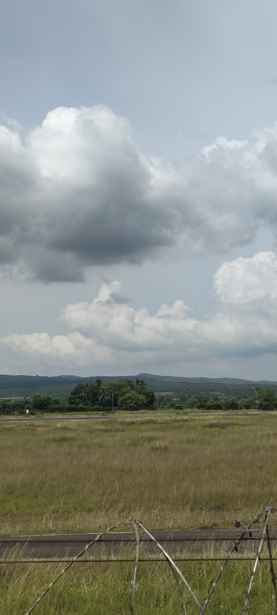 The image depicts a wide, open landscape under a cloudy sky. In the foreground, a grassy field stretches towards a road. A barbed wire fence is visible at the very bottom of the frame, its structure slightly out of focus. Beyond the road, a dense cluster of trees forms a prominent green island in the middle distance, with scattered palm trees interspersed among broader-leafed varieties. Further in the background, rolling hills covered in lush green vegetation recede into the horizon. The sky is filled with large, dramatic clouds, some appearing bright white and puffy, while others are darker and more ominous, suggesting an overcast or approaching storm. There are no people or animals visible in the scene. The overall impression is one of a rural or semi-rural environment, possibly on the outskirts of a town or city. This specific scene does not provide any unique identifying features that would pinpoint its exact location within Buengbakjok, Indonesia.