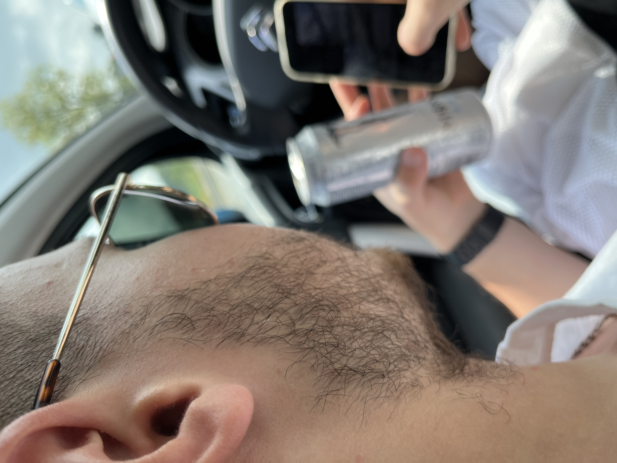 The image shows a close-up, angled view inside a car, with the head and upper body of a person in the foreground, largely out of focus. This individual appears to be resting or leaning back, with sunglasses perched on their head. The visible details include their ear, jawline, and short, dark hair or stubble.
In the blurred background, another person's hands are visible. One hand holds a smartphone, while the other grasps a silver beverage can. The can features blurred text that appears to be Japanese characters. A white, possibly mesh, fabric is seen around these hands, along with a black wristband or watch on one wrist. A portion of a car's steering wheel is also visible in the background, confirming the vehicle setting. Through the car window, a bright, possibly sunny, sky and blurred green foliage are discernible, indicating daytime.
The scene depicts a casual moment inside a car, possibly during a journey or a break, with one person relaxing and another holding a phone and a drink. The location context provided is Ruen, Bulgaria.