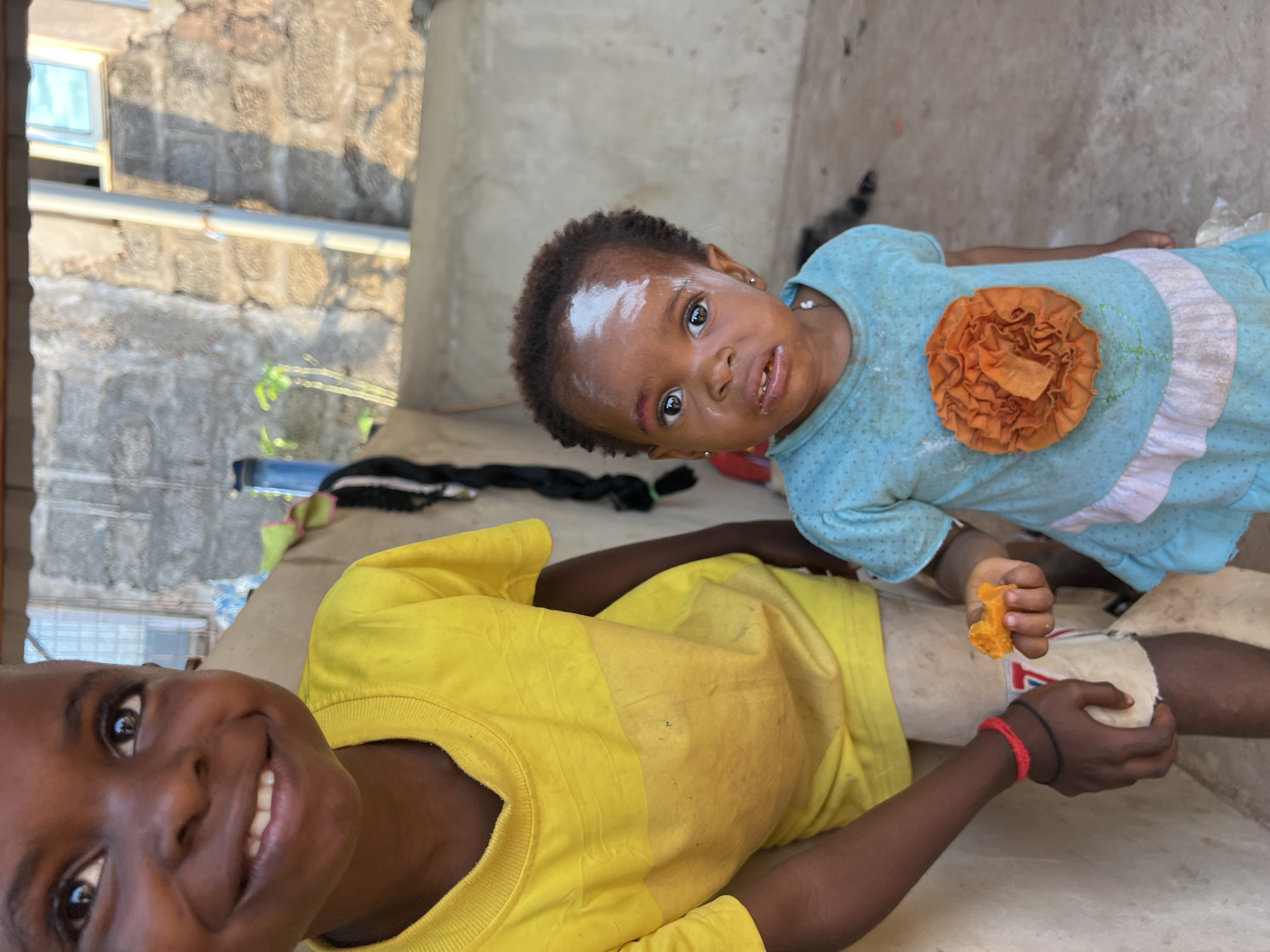 The image shows two young children, likely siblings, outdoors in what appears to be a residential area in Nkpor, Nigeria. The child in the foreground, facing the camera and wearing a bright yellow t-shirt, has a broad smile and a joyful expression. Their face is framed by dark hair. Behind them, a younger child is lying down, looking directly at the camera with wide, curious eyes. This child is wearing a light blue dress with a decorative orange flower applique on the chest and a white ruffled trim. They have a white substance applied to their forehead and cheeks, possibly traditional adornment or face paint.
The ground beneath them is a concrete or dirt surface, with some debris visible. In the background, a rough, textured stone wall with a window opening is discernible, suggesting a simple, possibly rural or semi-urban dwelling. A white pipe runs horizontally across the scene in the background. The lighting suggests it is daytime, with shadows indicating the sun's position. The overall mood is one of happy, everyday life. No visible text is present in the image.