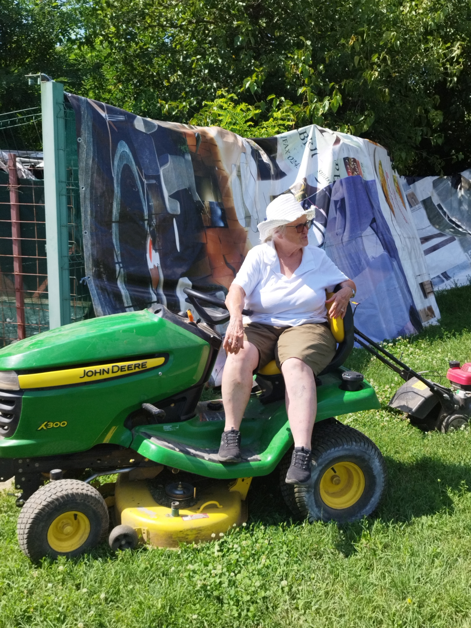 An elderly Caucasian woman wearing a white sun hat, sunglasses, a white polo shirt, and khaki shorts is sitting on a green John Deere X300 lawn tractor. The tractor is parked on a grassy lawn in what appears to be a residential backyard. Behind the tractor is a tall fence with a metal mesh and a large tarp or banner hanging from it. The banner displays a collage of images, some of which appear to be advertisements or posters. The woman is looking to her right, away from the camera. A smaller, red and black lawn mower is parked to the right of the tractor. The scene is illuminated by bright sunlight, suggesting it is daytime. The background is filled with lush green trees and foliage. The location is the city of Bucharest, Romania.