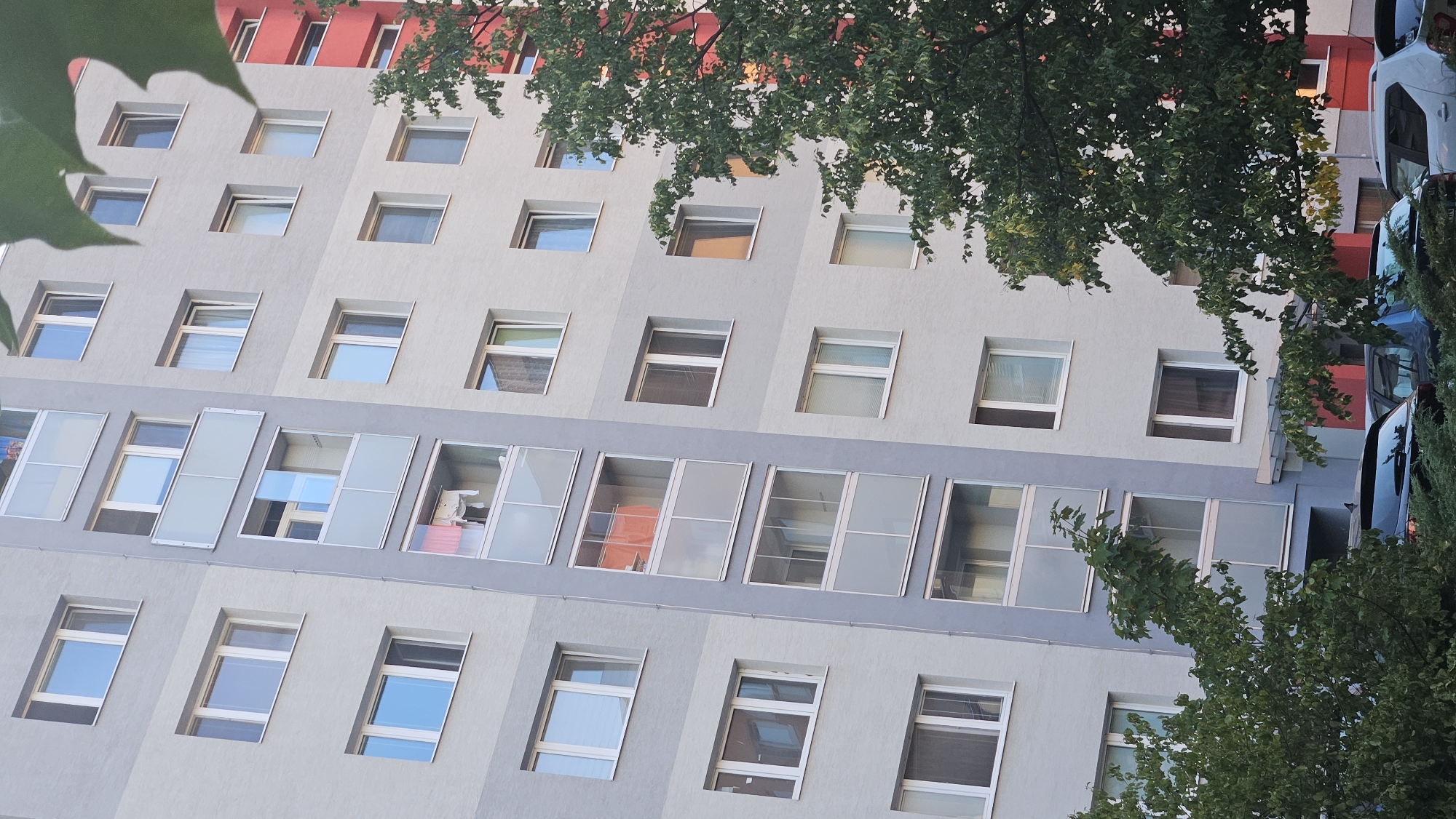 The image depicts a modern apartment building in Bratislava, Slovakia. The building features a grey facade with numerous windows and balconies. One vertical section of the building is lighter grey and incorporates glass-enclosed balconies, some of which show laundry hanging out to dry and an orange fabric. To the right of the grey building, a section of a red apartment building is visible. The foreground is partially obscured by lush green trees and bushes, with several cars parked in front of the building, including a black SUV and a silver car. The sky is not visible in the frame.