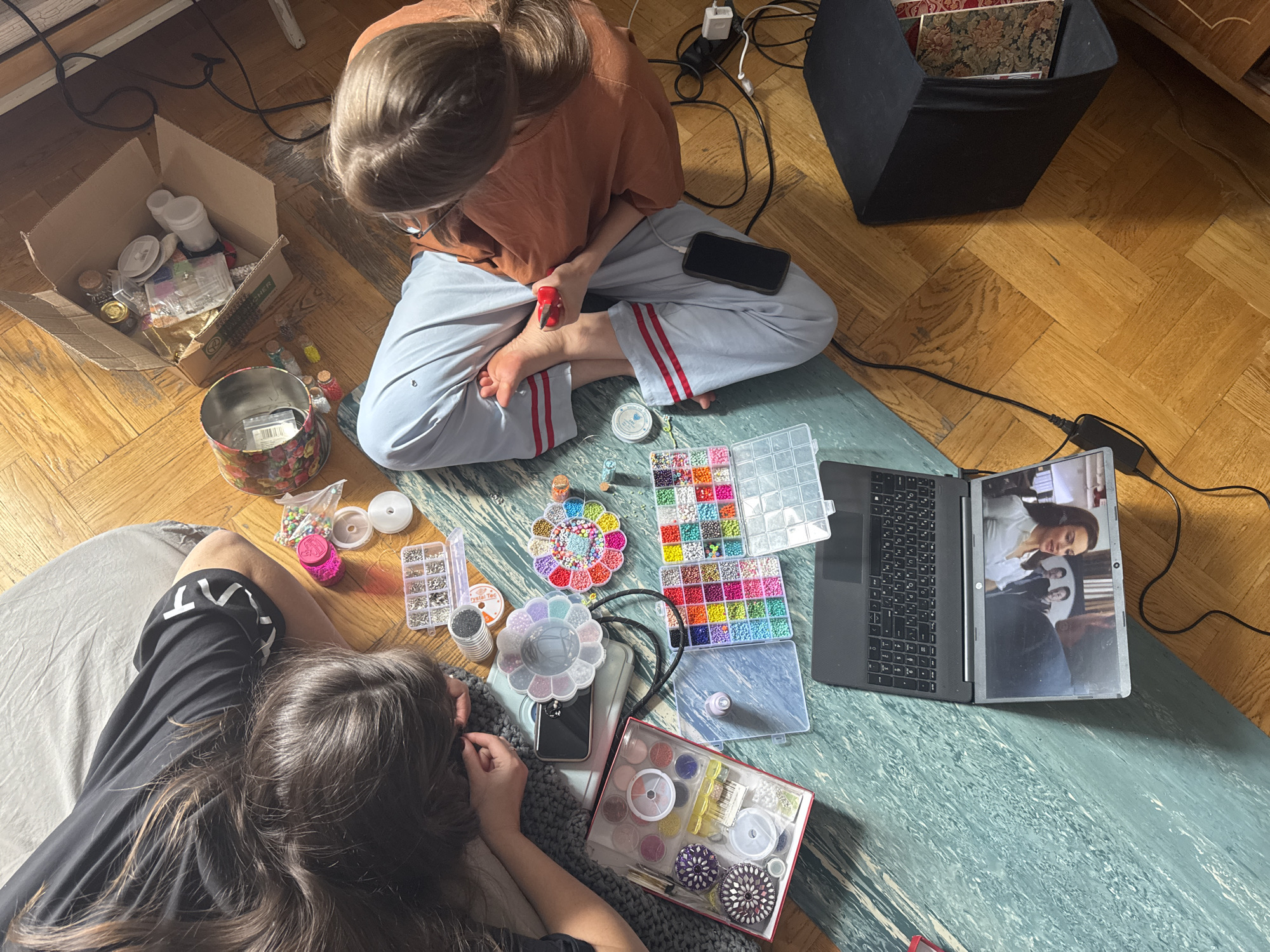 Two people sit on a blue and green patterned rug on a parquet floor. One person, seen from above, wears an orange shirt and grey pants with red stripes. The other person, partially visible, wears a black shirt. They are surrounded by crafting materials, including boxes of beads, containers, and a laptop. The laptop screen displays a video of a woman in a white shirt and other people. A black box and a cardboard box are also visible in the background. Electrical cords and wires are scattered near the box. The location cannot be verified as Moscow, Russia, from the image.