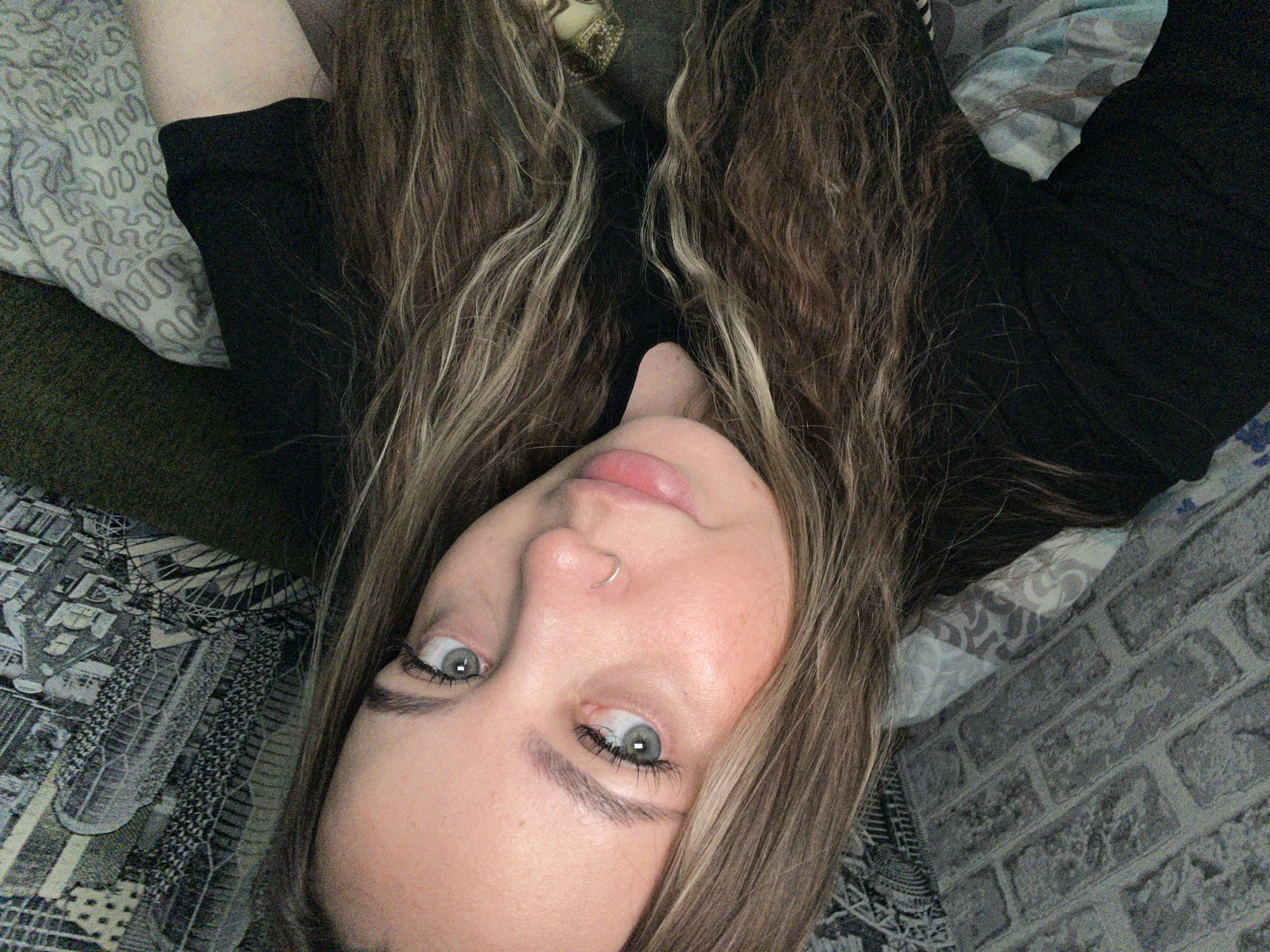 A young woman with long, brown and blonde hair, grey eyes, and a nose ring is lying down. She is wearing a black shirt. She is lying on a bed with different patterned bed sheets.