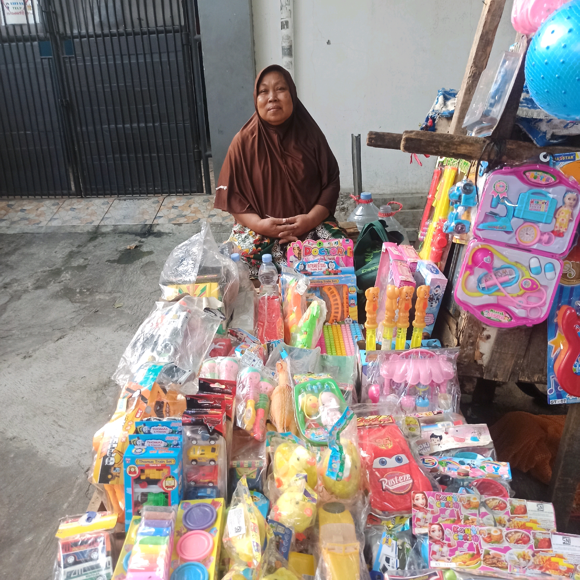 A street vendor, a woman wearing a brown hijab, sits behind a colorful display of toys. The toys, some still in their packaging, include miniature vehicles, play doctor kits, toy figures, and bubble wands. Behind her, a black metal gate suggests the location is a storefront or an entrance. The ground is a concrete pavement with scattered debris. The scene is set outdoors, likely in a bustling urban environment, possibly a market or a sidewalk, in Central Jakarta, Indonesia.