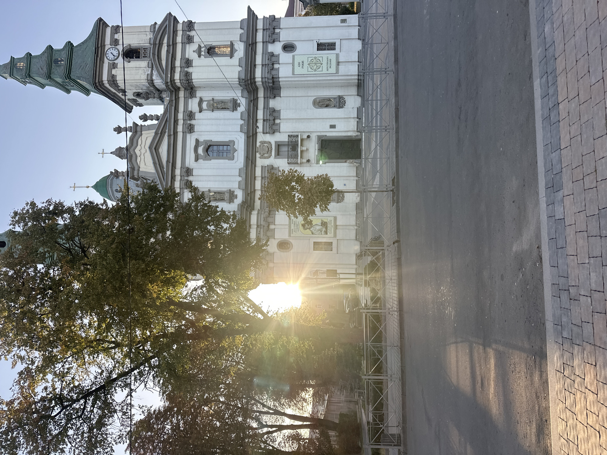 The image shows a wide, sun-drenched view of the Ternopil Castle Church of the Immaculate Conception, located in Ternopil, Ukraine. The grand, white baroque-style building features a tall, green-roofed bell tower with a clock face. The sun's rays are streaming intensely from behind a large, leafy tree on the left, creating a lens flare effect and casting long shadows across the asphalt road and paved sidewalk in the foreground. A metal fence separates the sidewalk from a grassy area in front of the church. Several windows and architectural details are visible on the church's facade, including statues and decorative elements. The sky is clear and blue, indicating a sunny day. There are no people clearly visible in the scene.
