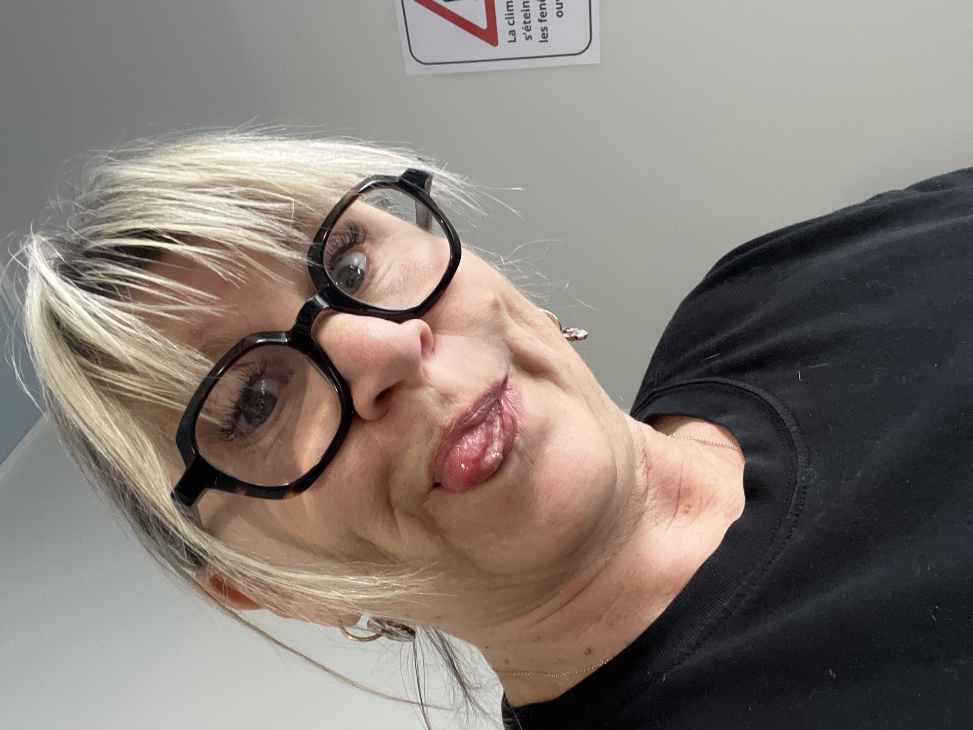 A woman with short, blonde hair and black-rimmed glasses is seen from a low angle. She is wearing a black shirt and sticking her tongue out slightly. A silver chain necklace is visible around her neck. In the top portion of the image, there is a sign in French; “La clim s’éteint les fenêtres ouvertes.” The image was taken indoors, likely in Saint-Galmier, France.