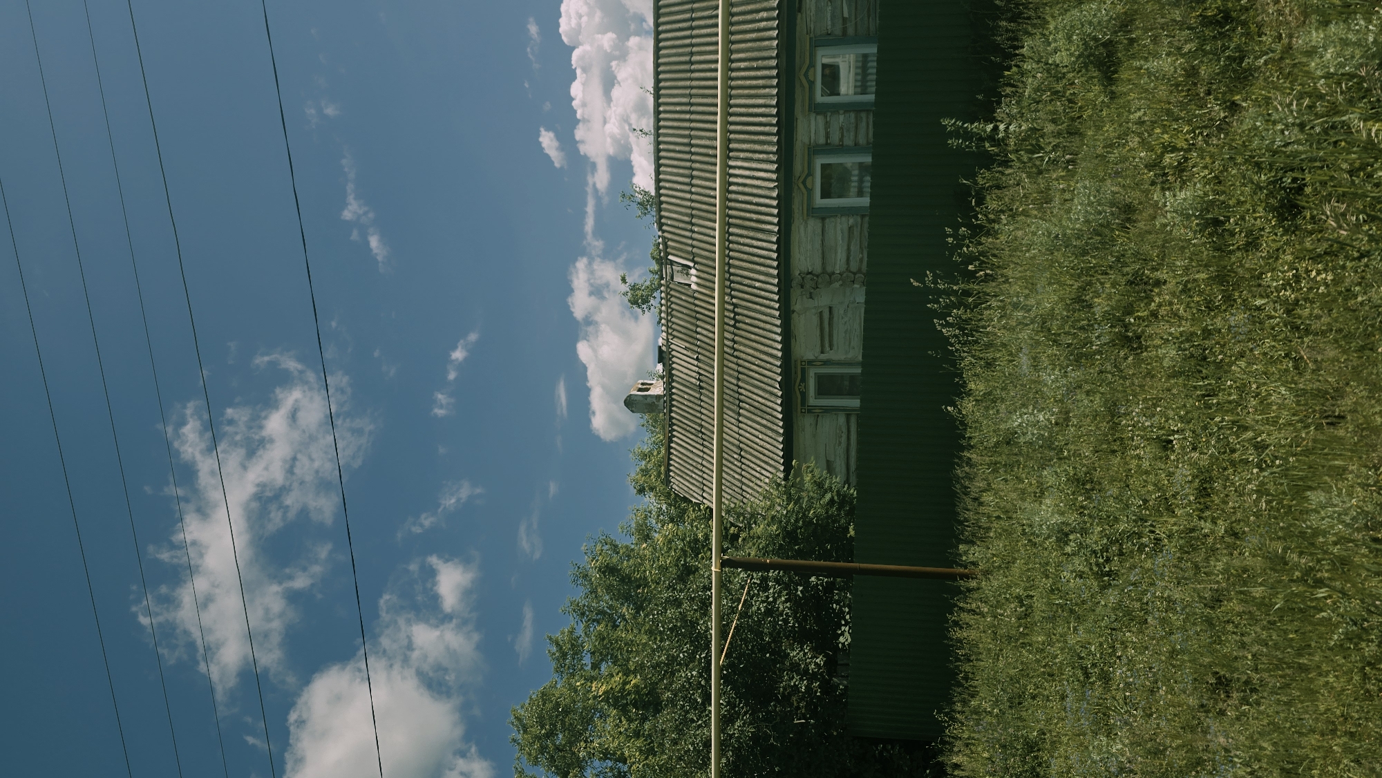In Kzyl-Chishma, Russia, a weathered, wooden house with a corrugated roof stands behind a dark green metal fence. The house features three windows with decorative trim and a stone chimney on the roof. Tall green trees surround the house, and a yellow pipe runs horizontally in front of it, supported by a brown pole. The foreground is filled with a dense field of tall, green grass and wildflowers. The sky above is a clear blue, dotted with fluffy white clouds, and several dark power lines stretch across it.