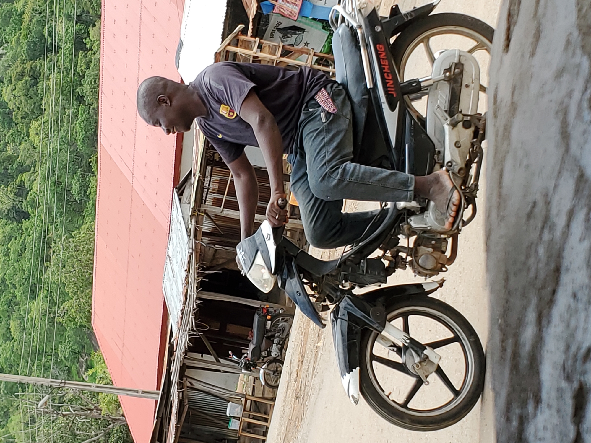 A man is depicted on a motorcycle in Mararaba, Nigeria. The man is Black, wearing a dark T-shirt, jeans, and sandals. He is bent over the motorcycle's handlebars. The motorcycle is black and silver, with the word "JINCHENG" visible on the rear. The background includes a building with a red roof and a hillside with trees. Another motorcycle is visible in the background. The scene is set on a road in daylight.