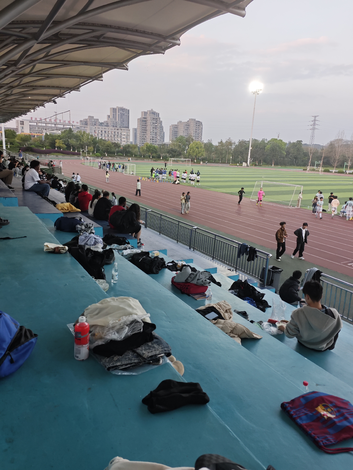 The image displays an outdoor sports stadium at the Chongqing University Huxi Campus Sports Center, situated in the Chongqing High-tech Zone, China. The scene is observed from an elevated position within the spectator stands, under twilight or early evening conditions indicated by a brightly illuminated floodlight and a light grey, overcast sky.
In the foreground, multiple young adults are seated on teal-colored tiered concrete stands. Numerous personal items are placed on the seats, including backpacks (one blue), various articles of clothing, clear plastic water bottles (one with a red cap), and food containers. A blue and red FC Barcelona drawstring bag rests on a lower step.
The midground features a reddish-brown synthetic running track encircling a green artificial turf soccer field. Several groups of individuals, predominantly young adults, are present on the track and field. Some are wearing athletic attire, including blue and white jerseys, while one individual near a soccer goal is dressed in a bright pink full-body suit. Others are casually dressed. Actions observed include standing in groups, walking, and observing. A line of orange cones is visible on the field. Two white soccer goals are positioned at opposite ends of the field.
The background reveals a sequence of multi-story buildings, likely residential or institutional, rising above a line of trees beyond the sports complex. A tall floodlight tower illuminates the field from the right side of the image, and a high-voltage power transmission tower is visible further in the distance. The upper portion of the stadium's structure, a large, curved tensile fabric roof supported by metal trusses, spans across the top of the frame.