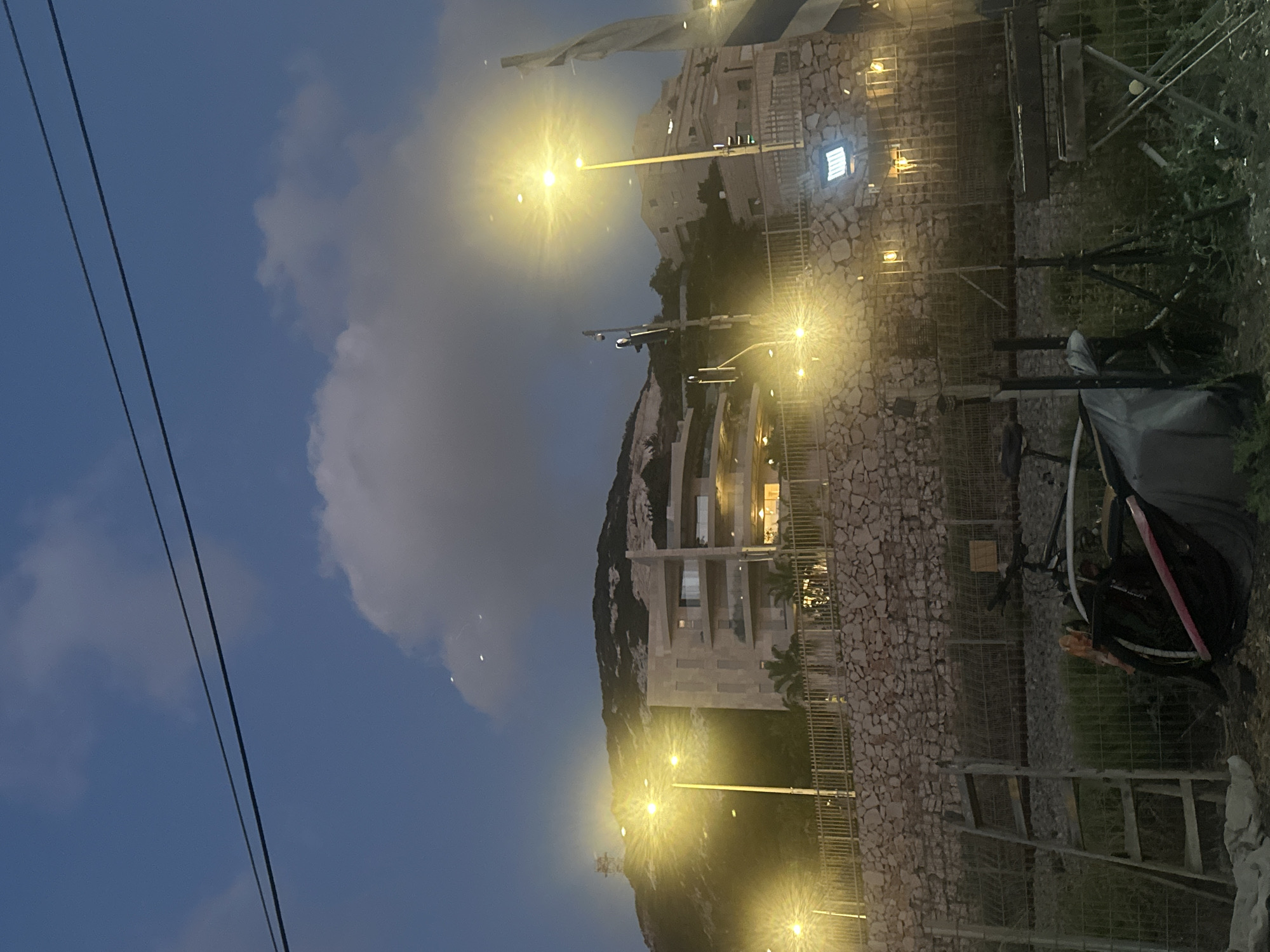 The image depicts a nighttime scene in Haifa, Israel, with a modern building constructed on a rocky hillside. The sky is dark blue with scattered clouds, and streetlights illuminate the area, casting a warm glow. The building appears to be multi-story with visible windows, some of which are lit from within. A stone wall with a metal fence runs along the foreground, separating the property from the street.
To the right, there are several objects, including a ladder leaning against the fence and what appears to be a bicycle or motorcycle with a black cover. The overall mood is calm and tranquil, characteristic of a quiet evening in a city. The presence of streetlights suggests the scene is taking place after sunset, likely in the early evening or night. The rocky terrain and architecture are consistent with the landscape of Haifa, a city known for its hilly terrain and coastal views. No people are visible in the image, and there is no discernible text.