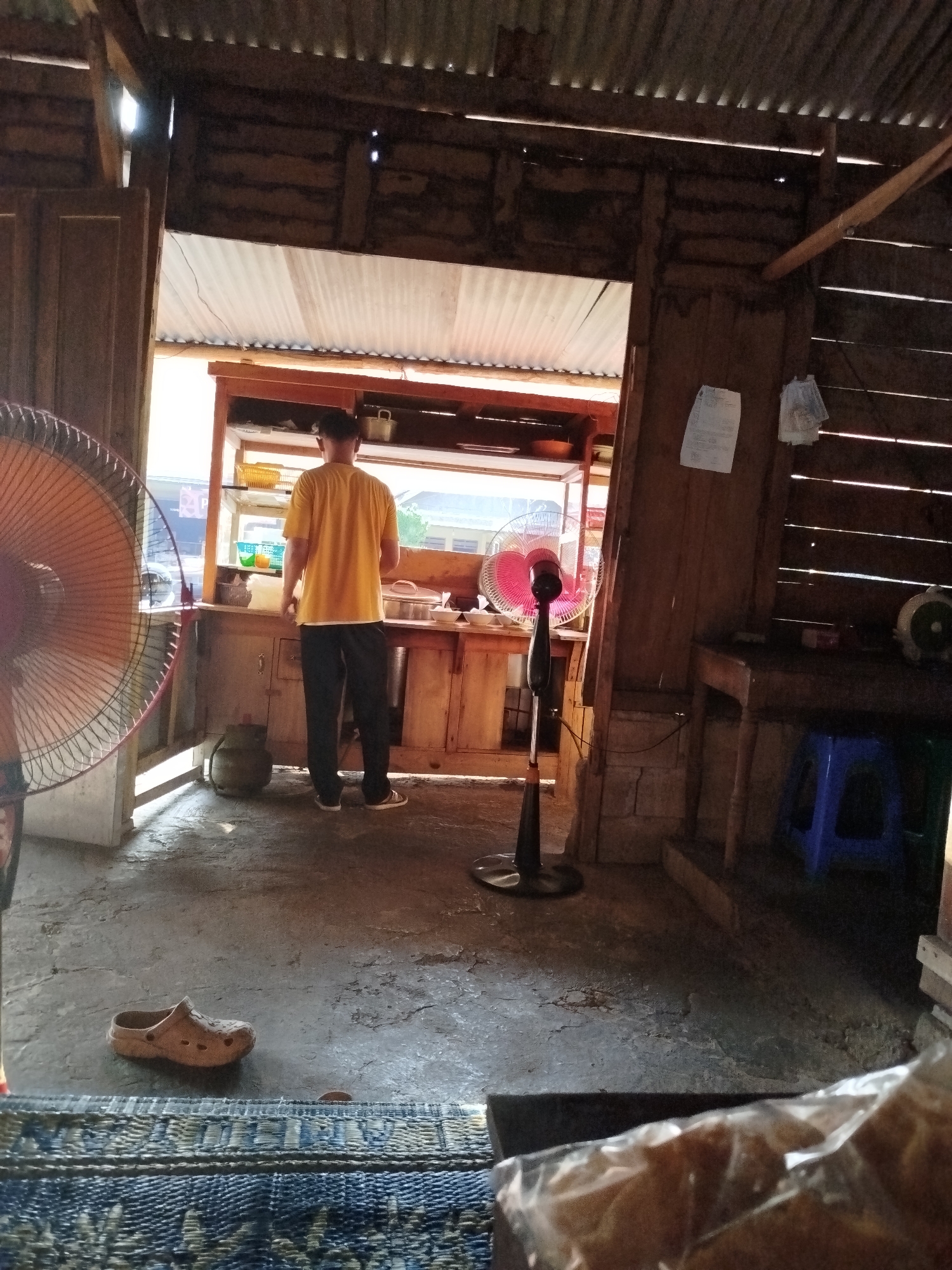 The image depicts an indoor scene, likely a food stall or small eatery, situated in Pojok, Indonesia. A person wearing a yellow t-shirt and black pants stands facing away from the camera, positioned behind a wooden counter. The counter displays various items, including bowls, a gas cylinder, and what appears to be food preparation equipment. To the left of the person, a large orange fan is visible, and a smaller black oscillating fan stands in front of the counter. The stall has a rustic, open-air construction, with wooden walls and a corrugated metal roof, allowing natural light to enter. In the foreground, a single brown clog is on the floor, and a portion of a woven mat is visible. The overall environment suggests a simple, functional establishment.