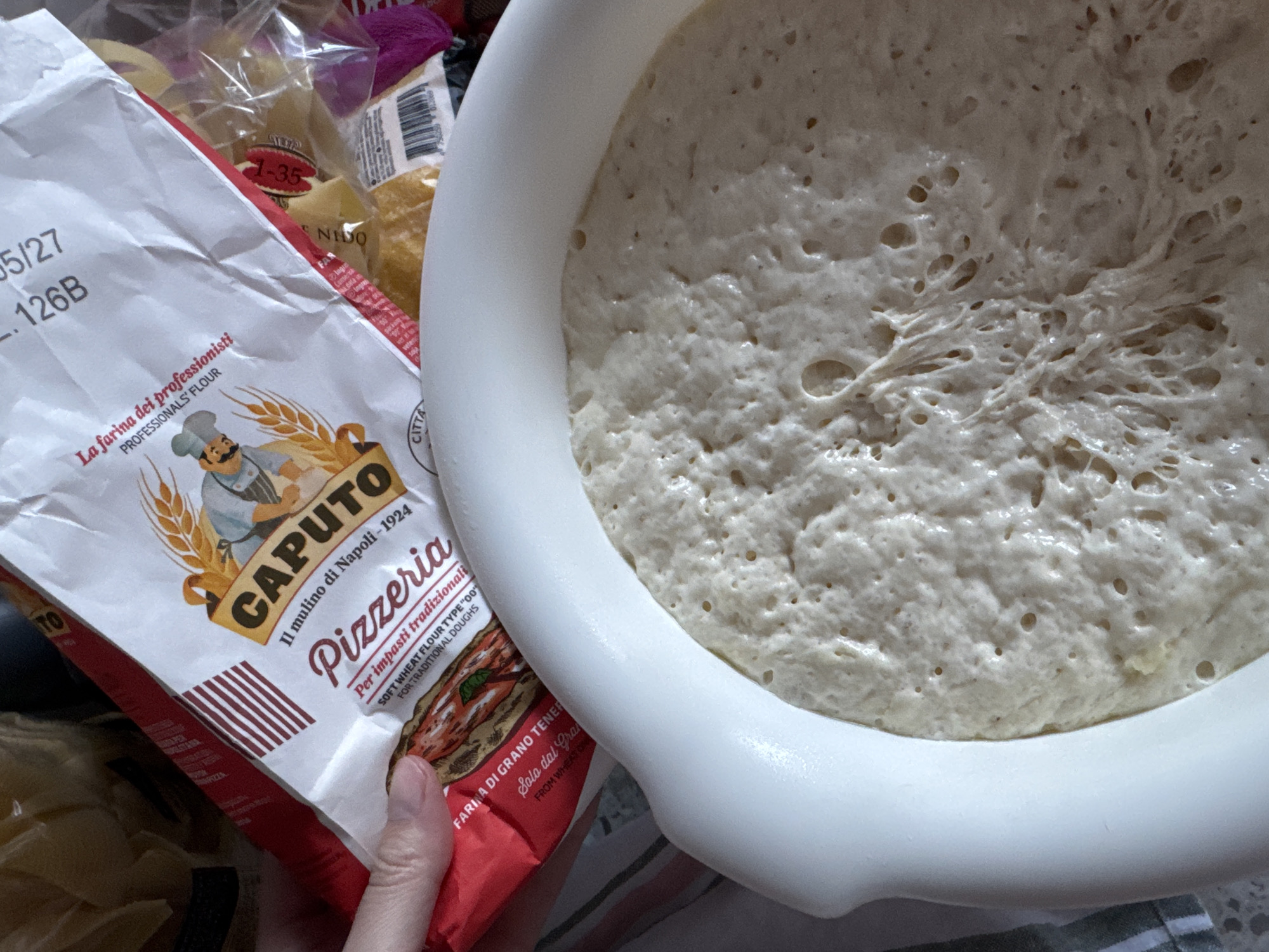 The image shows a white bowl filled with bubbly, risen dough next to a bag of Caputo Pizzeria flour. The flour bag is red and white, with an image of a chef and wheat on the front. The flour is labeled as "Il mulino di Napoli-1924," indicating it's from Naples, Italy. There's pasta visible in the background, along with a partially visible hand holding the flour bag. The scene appears to be set indoors, likely in a kitchen. There is no evidence in the photo to confirm the city of Riga, Latvia.