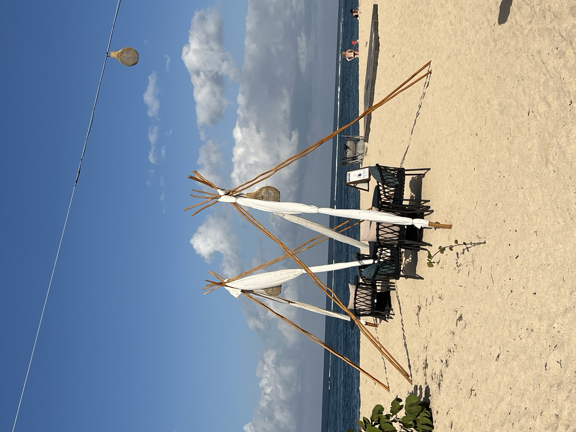 The image depicts a tranquil beach scene in Kuta Selatan, Indonesia, bathed in bright daylight under a clear blue sky with scattered fluffy white clouds. The foreground is dominated by a sandy beach, with the gentle waves of the ocean visible in the background. Two picnic-style seating areas, constructed from bamboo poles and draped with white fabric, are set up on the sand. Each seating area features a table surrounded by dark wicker chairs, some of which have light-colored cushions. A small green plant grows on the sand in front of the left seating area, and another tiny green sprout is visible in the sand between the two structures.
In the mid-ground, to the right of the seating areas, a wooden platform is laid out on the sand, and several people are visible enjoying the beach. A man in red shorts stands on the platform, and another person is seated to his right. Further in the distance, other beachgoers can be seen. A white string stretches across the top of the frame, with a decorative, circular object resembling a woven basket or lamp hanging from it. A small, rectangular sign with the word "BEWARE" in white text is placed on the sand near the left seating area. The overall atmosphere is serene and inviting, suggesting a relaxing day at the beach.