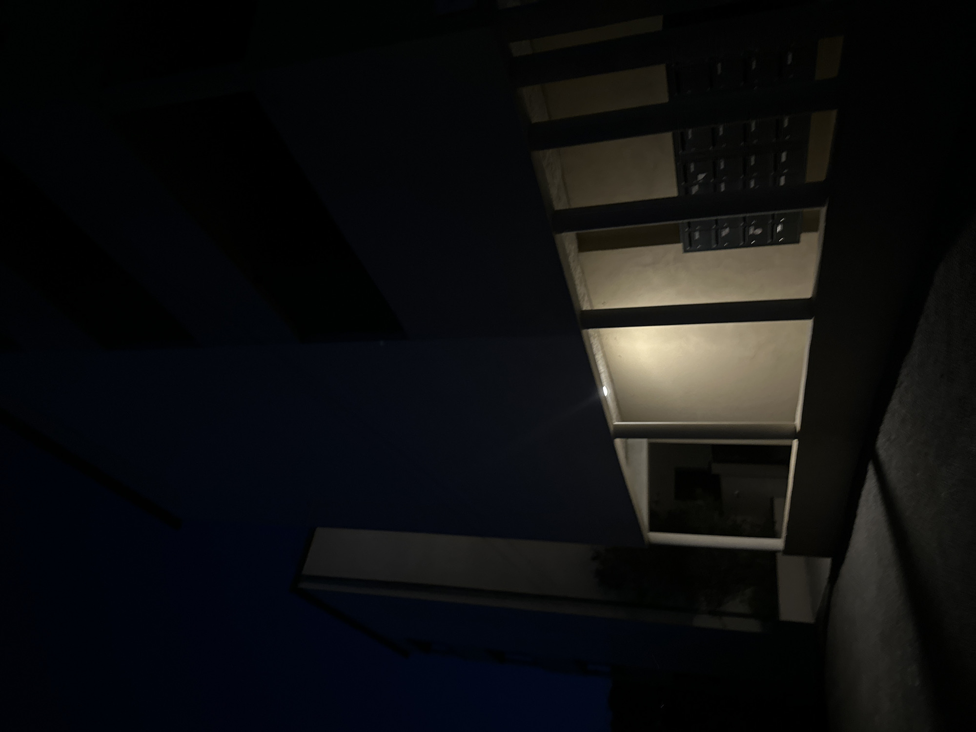 The image depicts the exterior of a modern building at night, likely in the city of Anduze, France. A tall, rectangular structure with a white facade is visible on the left. To the right, a darker building section recedes into the darkness. In the foreground, a walkway or patio area is illuminated by soft light emanating from within a sheltered section of the building. This sheltered area is characterized by a light-colored wall and a series of vertical support beams. Mounted on the wall are rows of dark, compartmentalized mailboxes, suggesting this is an entrance or common area. The ground in front of the building is dark and appears to be asphalt or concrete, with shadows cast from the structure. The sky above is a deep, dark blue, indicative of nighttime. There are no people visible in the scene.