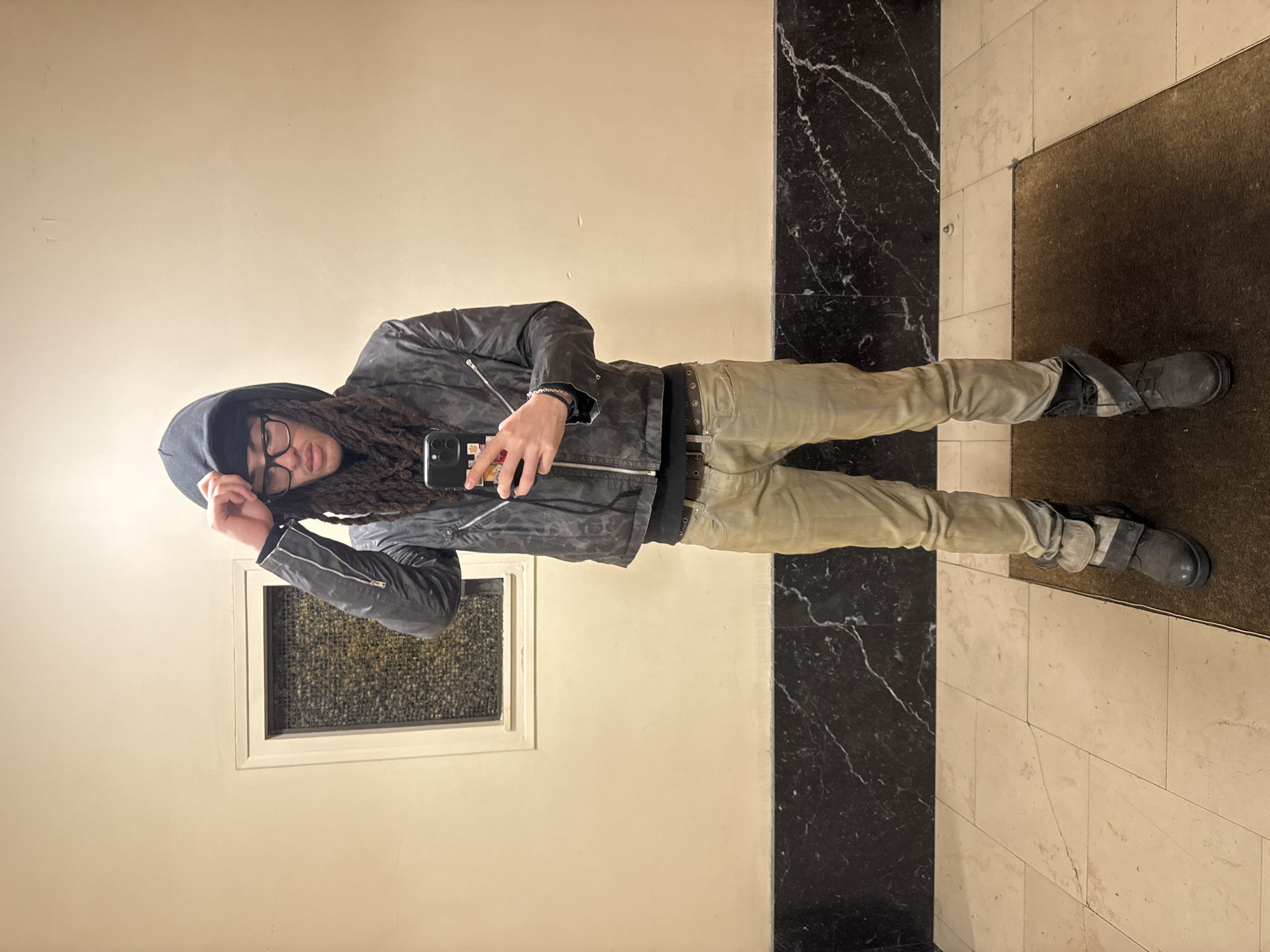 An individual stands facing the camera, likely capturing a mirror selfie in an indoor setting. The person has dark dreadlocks, wears clear-framed glasses, a black hooded top with the hood up, a dark gray bomber-style jacket with a subtle pattern and prominent zippers, light khaki-colored pants, a dark belt with eyelets, and dark grey high-top boots. A dark-cased smartphone with a multi-lens camera is held in the right hand, angled towards the viewer, while the left hand touches the hood. The background consists of a cream-colored wall, featuring a rectangular recessed window or panel with textured glass on the left. A horizontal band of dark, veined marble lines the lower portion of the wall. The floor is composed of light-colored tiles, with a dark brown rectangular mat positioned beneath the individual's feet. No specific visual identifiers within the image confirm a precise location within the city of Paris, France.