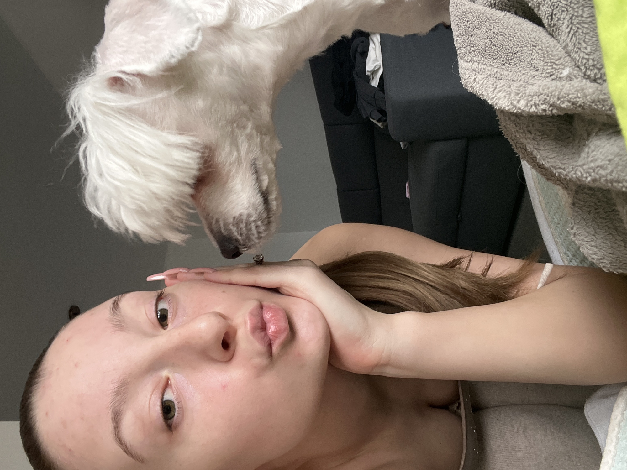 A woman and a dog are positioned in an indoor setting. The woman is lying down, with her face close to the camera, displaying a pouting expression. Her right hand is raised near her face, with fingernails painted pink. Her brown hair is partially visible. Above her, a small, white dog is positioned, facing downward with its nose close to the woman's face. The background features a dark-colored couch with some clothing items visible on it. A grey blanket and a pale green object are also present in the background. There is insufficient information to identify the location as Varna, Bulgaria.