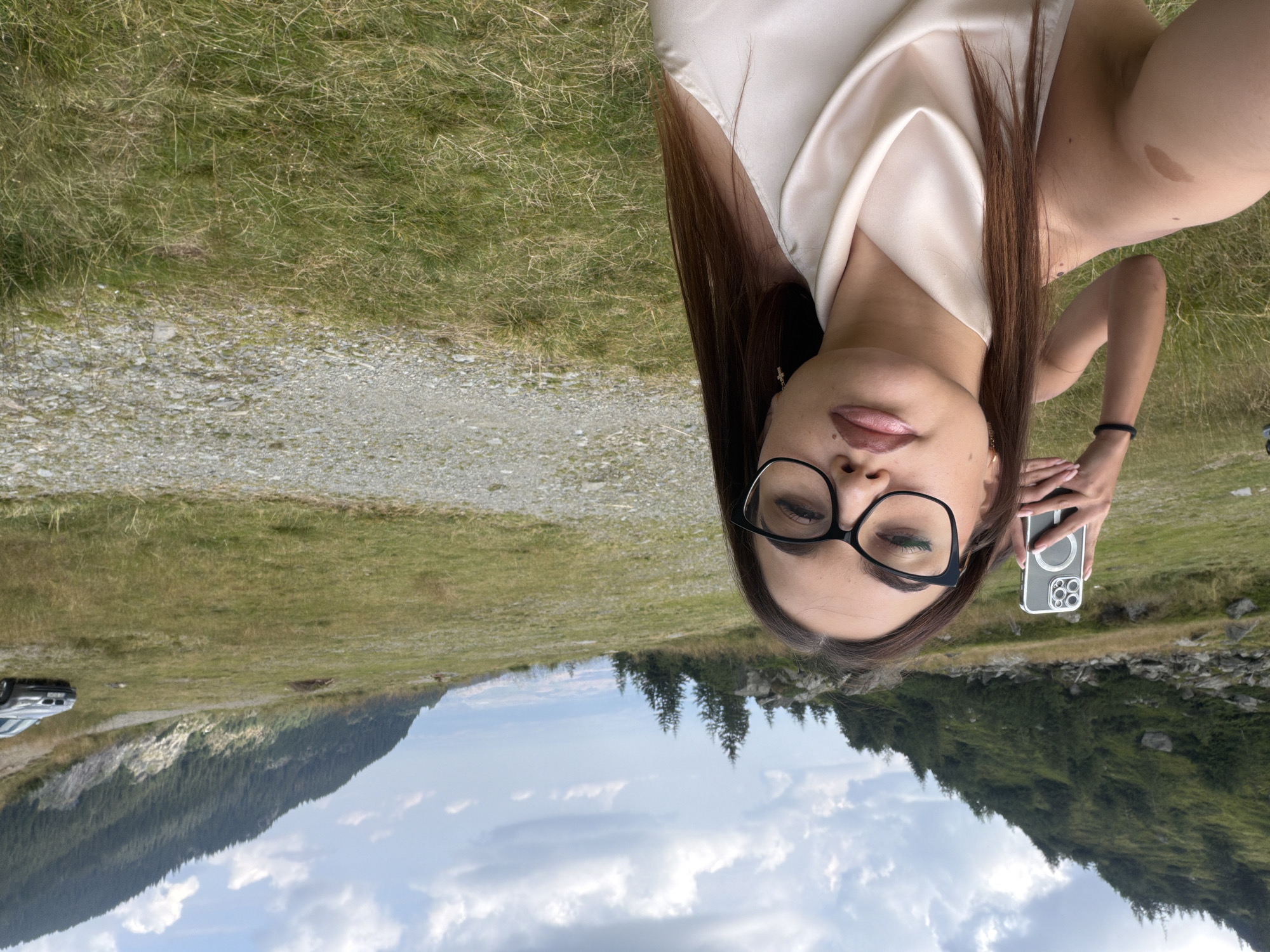 In the mountainous landscape near Cârțișoara, Romania, a young woman with long, dark hair and glasses is taking a selfie outdoors. She is wearing a light-colored, draped top. Behind her, another person is visible, holding up a smartphone, possibly to capture a wider shot or a different angle. The environment consists of a grassy slope with scattered rocks and a gravel path leading into the distance. Pine trees dot the hillside, and a silver car is parked on the grass in the background, suggesting a location accessible by road. The sky is partly cloudy, with dramatic cloud formations adding to the scenic vista.