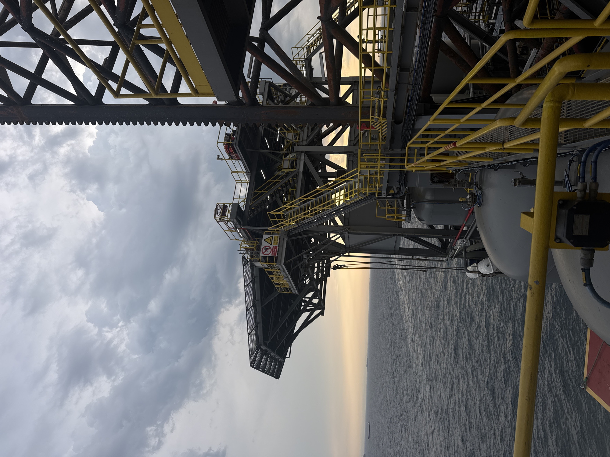 The image shows a portion of an offshore platform, likely a drilling rig, with a helideck extending out over the water. The sky is cloudy and overcast, with a hint of sunset glow near the horizon. The sea is calm with gentle ripples. Various metal structures, stairs, and yellow railings are visible, indicative of industrial infrastructure. A few distant ships can be seen on the water. The scene appears to be captured from another part of the same platform. No people are clearly visible in the image. The location is identified as Unknown, Italy, which is likely an error as offshore platforms are typically located in international waters or offshore the coast of a country.