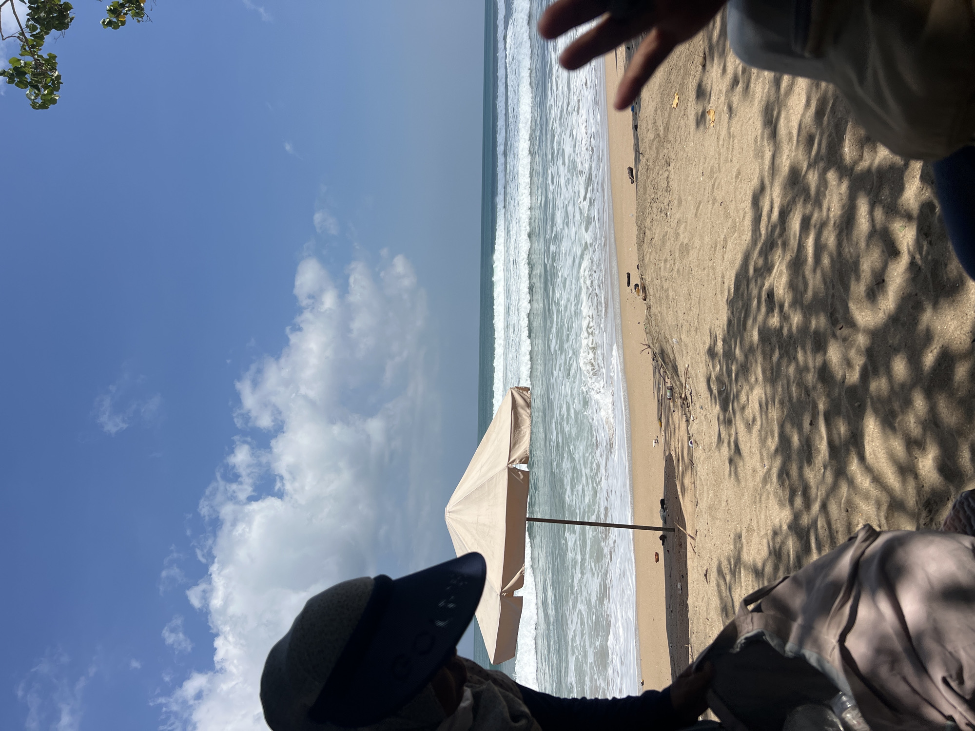 The image depicts a beach scene in Kuta, Indonesia, on a clear day with a blue sky dotted with white clouds. In the foreground, a person wearing a sun visor is partially visible on the left, with a bag next to them. In the middle ground, a light-colored beach umbrella stands on the sandy shore, facing the ocean. Gentle waves are rolling onto the beach. The background features the expansive ocean and horizon. The overall scene conveys a relaxed beach atmosphere.