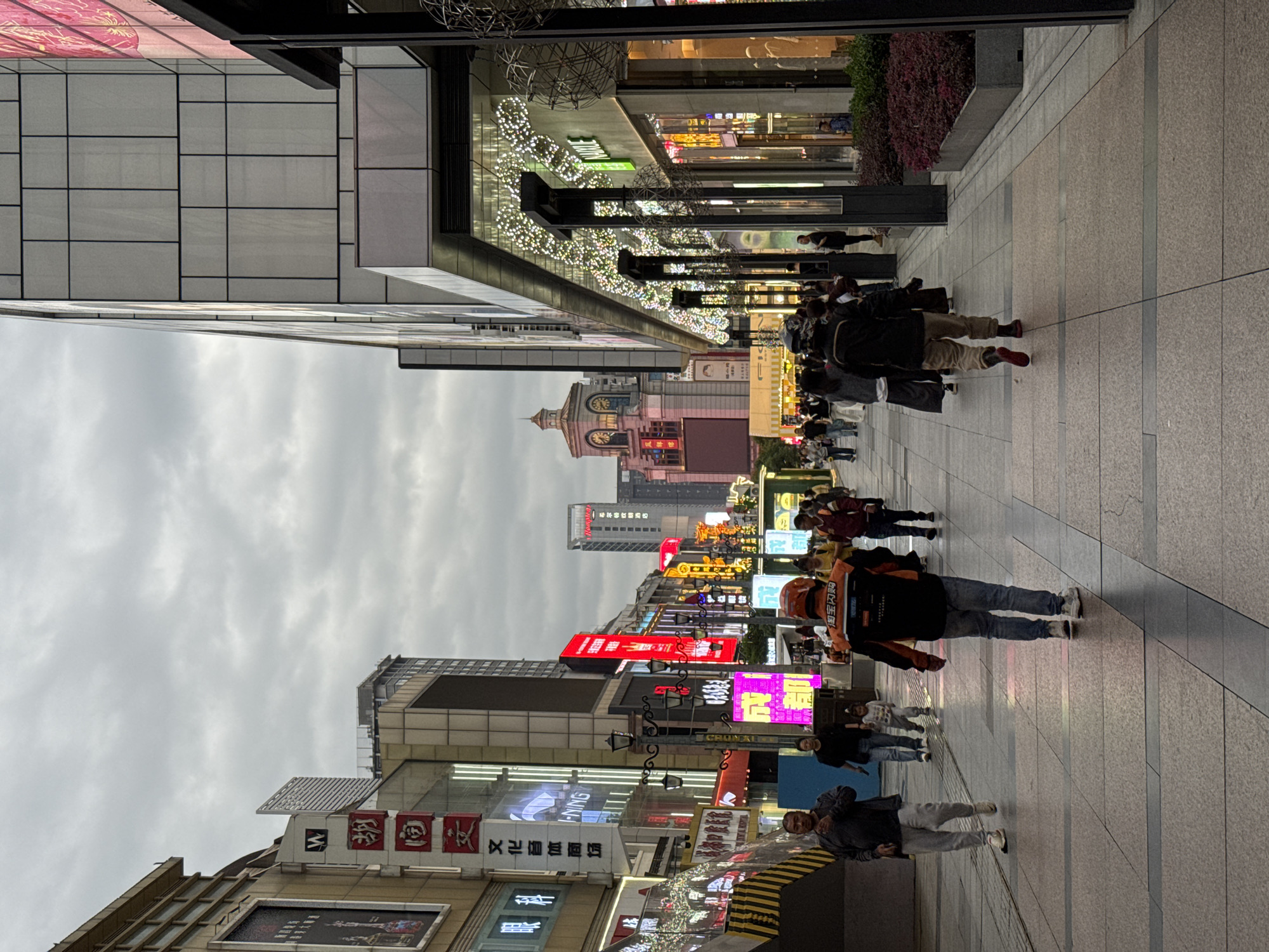 The image shows a busy street in Jinjiang District, Chengdu, China, lined with tall buildings adorned with bright neon signs and advertisements in Chinese characters. The sky is overcast. A diverse group of people walks along the wide, tiled pedestrian area. Some are carrying shopping bags or luggage. The buildings display a mix of architectural styles. A clock tower is visible in the distance. The street is decorated with festive lighting.