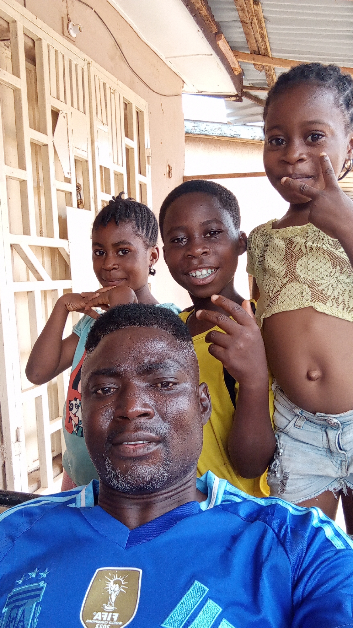 A man in a blue shirt with a FIFA logo is taking a selfie with three children. The children, two girls and one boy, are standing behind him, posing for the camera. The setting appears to be outside a house with a white grid pattern door. The image was captured in Sabon Tasha, Nigeria.