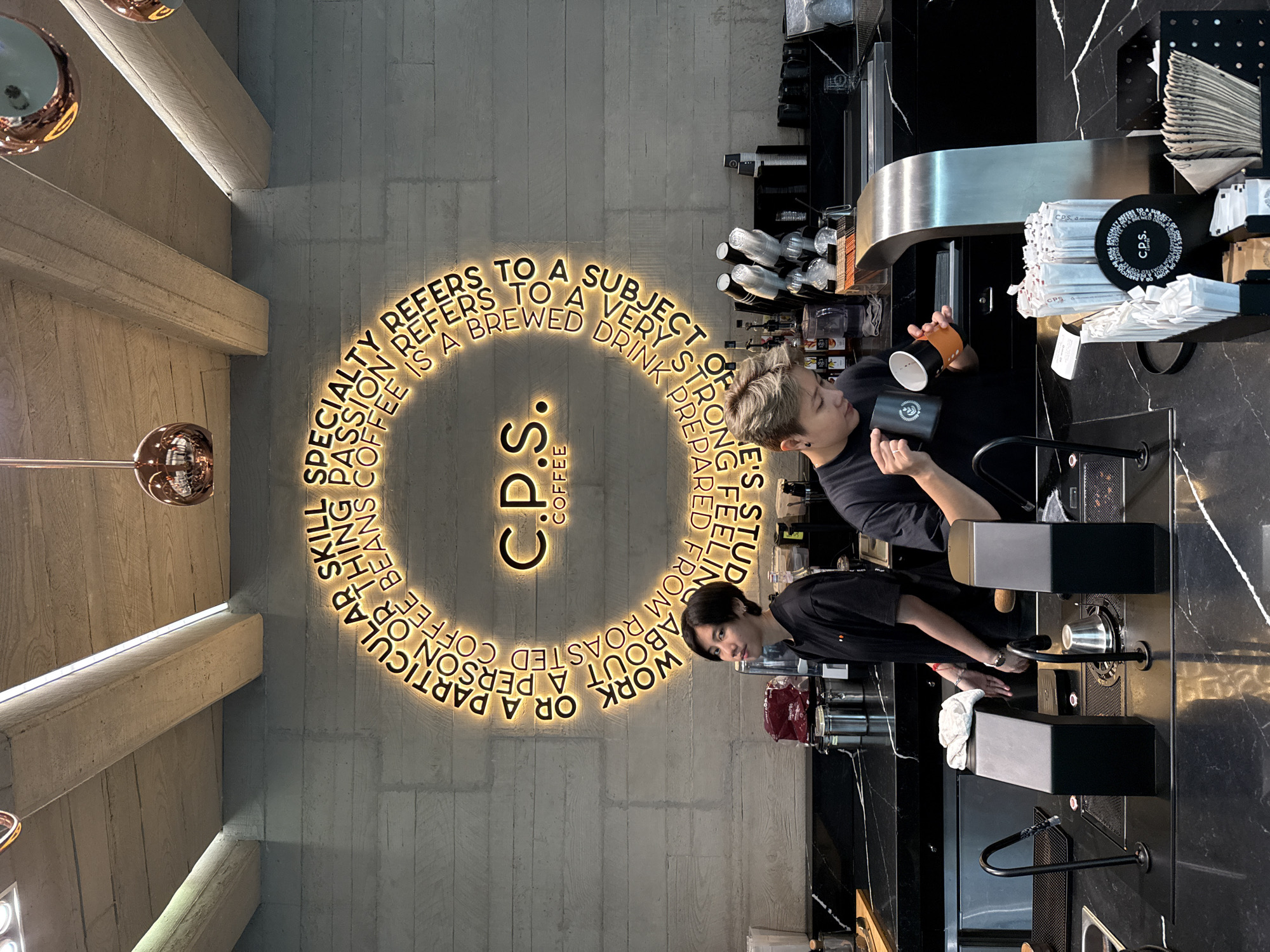 Here's a detailed description of the image:
**Environment:** The image is taken in a modern-looking coffee shop, possibly in Bangkok, Thailand. The interior design features gray walls with a circular neon sign that reads "CPS Coffee". Above, there's exposed beam construction with integrated lighting. The coffee bar area is sleek, with black marble countertops and stainless steel equipment.
**People:** The image shows two individuals, likely employees or baristas at the coffee shop. They are both wearing black t-shirts.
**Objects:** Key objects visible are coffee cups, coffee makers and related brewing equipment, drink stirrers, napkin holders, and what appear to be condiment containers.
**Actions:** The baristas appear to be in the process of preparing coffee. One is holding two cups, while the other is at a brewing station.
**Interactions:** The two individuals may be collaborating on the preparation of the beverage.
**Subject:** The image captures the atmosphere and workflow of a modern coffee shop, emphasizing the process of specialty coffee preparation.