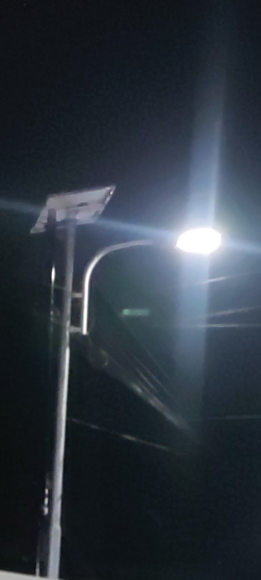 The image is a low-light, slightly blurred photograph of a solar-powered street lamp at night. The lamp post is metallic and vertical, with a solar panel mounted on top, angled slightly upwards. A curved arm extends from the post to hold the light fixture, which is brightly illuminated, casting a strong glare that obscures details. The background is dark and appears to be outdoors, with some faint horizontal lines possibly indicating overhead wires or distant structures. The scene is in the city of Ciledug, Indonesia. There are no people or specific actions depicted.