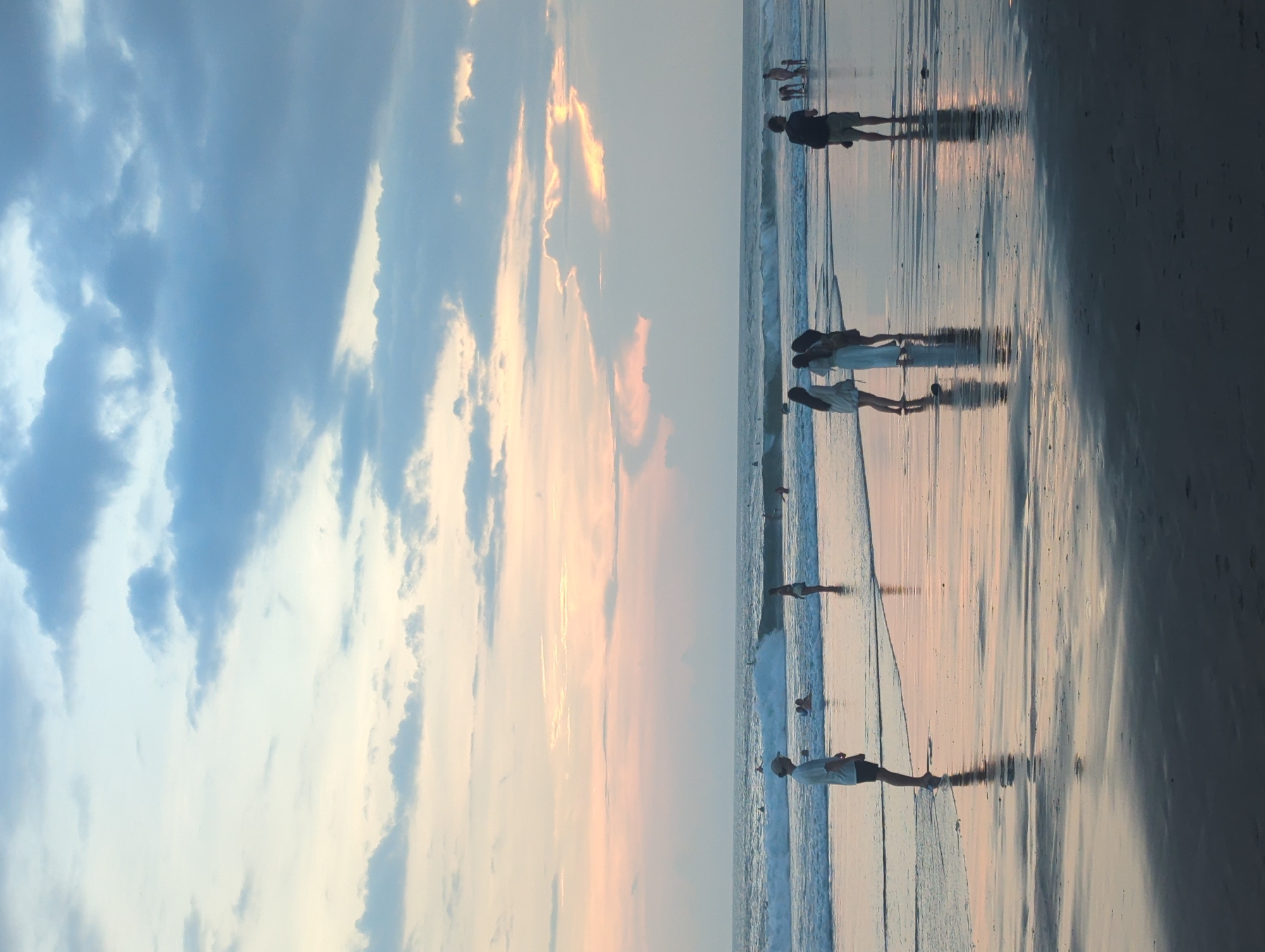The image depicts a beach scene at sunset in North Kuta, Indonesia. Several people are visible walking along the water's edge. The sky displays a gradient of colors, transitioning from blue to orange with the presence of clouds. The beach surface appears wet, reflecting the colors of the sky and the silhouettes of the people. Waves are gently breaking near the shore.