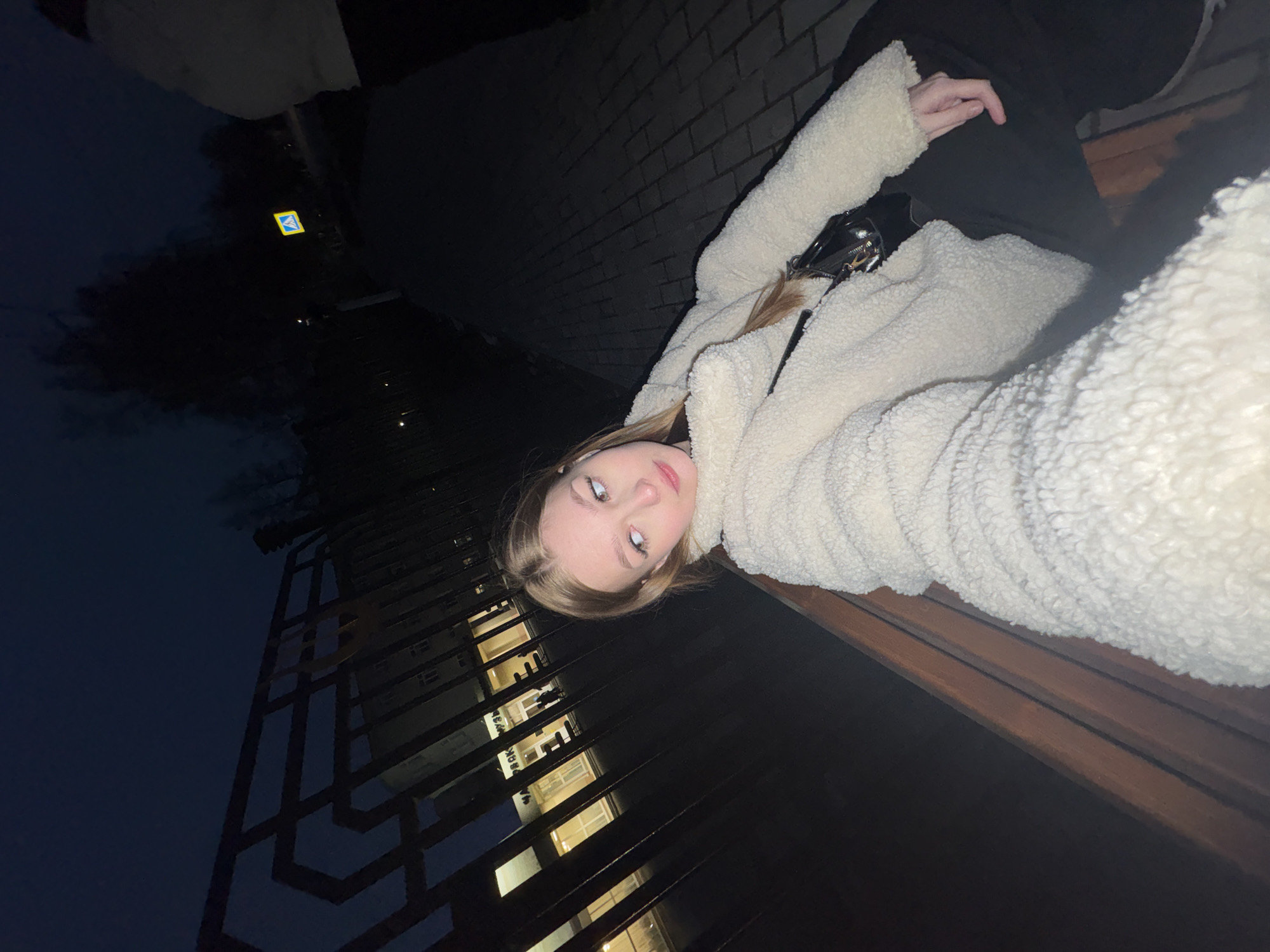 This media file captures a young woman lying horizontally, looking towards the camera, against an urban nighttime backdrop. She is wearing a cream-colored, fluffy jacket, possibly made of faux fur, and has light brown hair framing her face. A black shoulder bag with a metallic chain is visible near her chest. She appears to be resting on a dark wooden bench or ledge.
In the background, the sky is dark blue to black, indicating late evening or night. To the upper left, a bright yellow and blue square sign, characteristic of a pedestrian crossing warning, stands out against the darkness. Behind the woman, a textured dark brick wall is visible. To the lower left, a multi-story building with numerous illuminated rectangular windows can be seen through a dark, geometrically patterned metal grating or fence. Another person's leg and hand, clad in black, are partially visible in the upper right corner, suggesting companionship or a relaxed pose. The lighting is low, relying on artificial sources from the building and the sign. No explicit readable text is visible apart from the pedestrian crossing symbol. The image is composed with a strong diagonal line, emphasizing the woman's pose and the surrounding elements of the Chaikovsky, Russia setting.