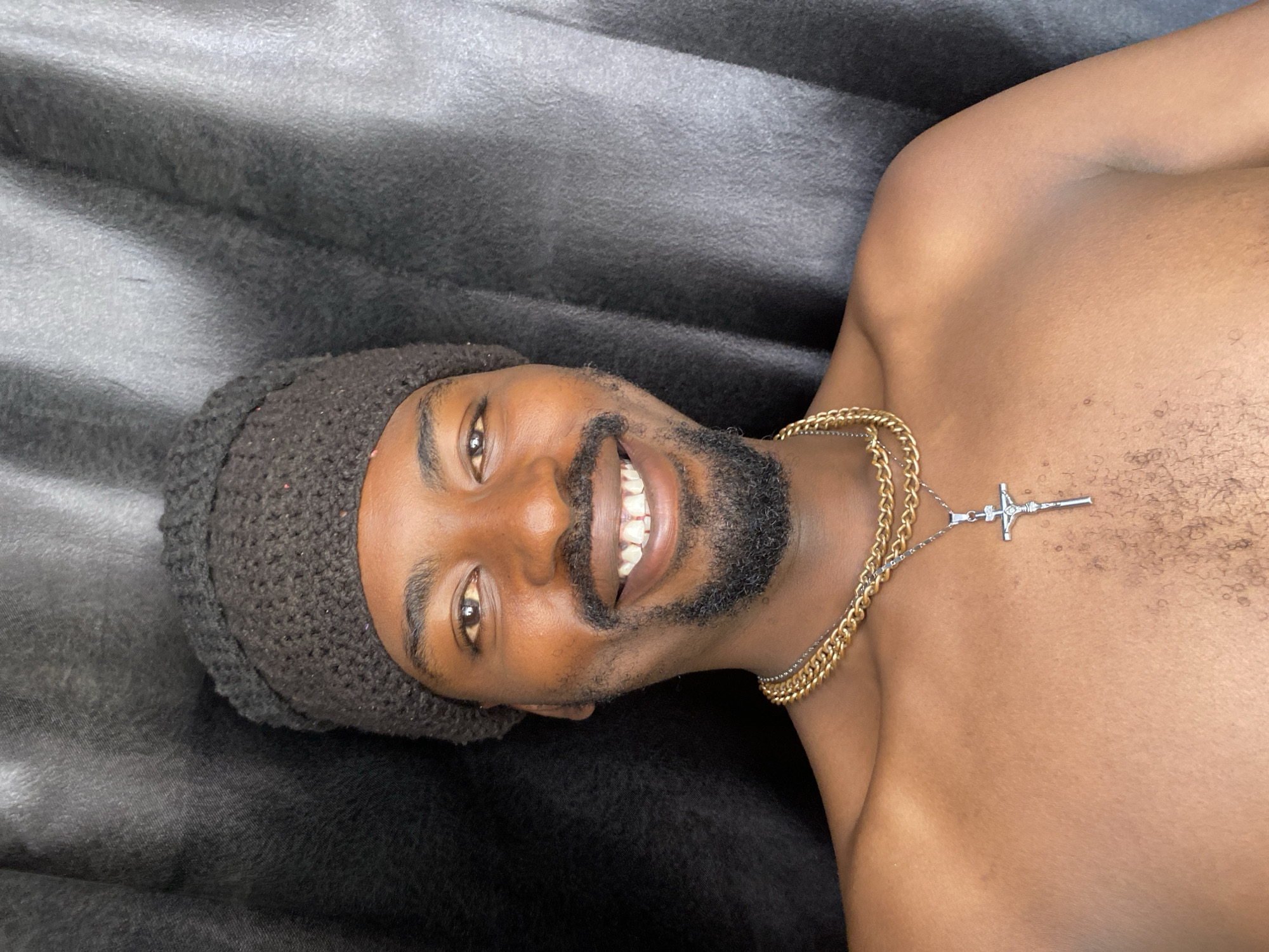 A dark-skinned man is captured in a close-up, smiling broadly at the camera. He has warm brown eyes, a full dark beard, and a mustache. He is wearing a dark, possibly knitted, beanie on his head. His wide smile reveals prominent white teeth, conveying a happy and friendly expression. He is shirtless, showcasing his chest and shoulders, which have some visible body hair. Around his neck, he wears two necklaces: a thick, gold-colored chain and a thinner, silver-colored chain adorned with a silver crucifix pendant.
The man is lying on a dark gray or black textured fabric, which appears soft, like a blanket or duvet, exhibiting subtle folds and varying light reflections. The lighting is soft, illuminating his face and upper body. The image is oriented as if the man is lying on his left side, with his head on the left and body extending to the right. There is no visible text within the image. This scene takes place in Barakin Akawo, Nigeria.