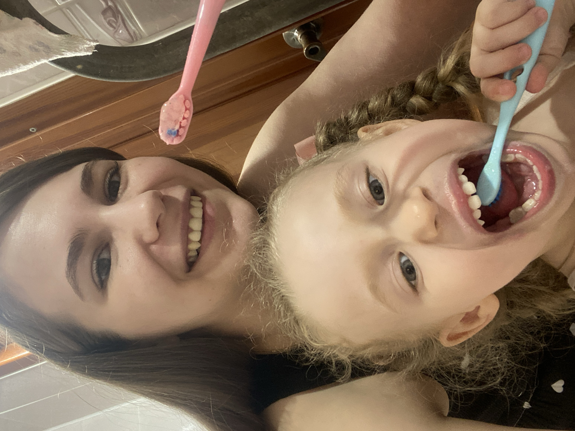 A close-up indoor photograph depicts two females in a domestic bathroom setting. An adult female, with long brown hair, occupies the upper portion of the frame, smiling directly at the camera. She is wearing a dark top. Her right hand, visible in the upper right quadrant, holds a pink toothbrush with pink toothpaste, which includes a distinct blue speck.
A younger female, a child estimated to be between three and five years old, is positioned in front of and below the adult. She has blonde, curly hair styled into at least one visible braid on the right side and wears a pink bow on the left side of her head. The child is looking towards the camera with her mouth wide open, actively brushing her teeth with a light blue toothbrush held in her right hand. Blue toothpaste is visible on the brush and in her mouth.
The background on the right side of the image features wooden paneling or a wooden door, on which a metallic door handle or knob is visible. A piece of white, patterned fabric or a towel is partially visible in the upper right. The specific location within Chaikovsky, Russia, is not discernible from the visual evidence provided.