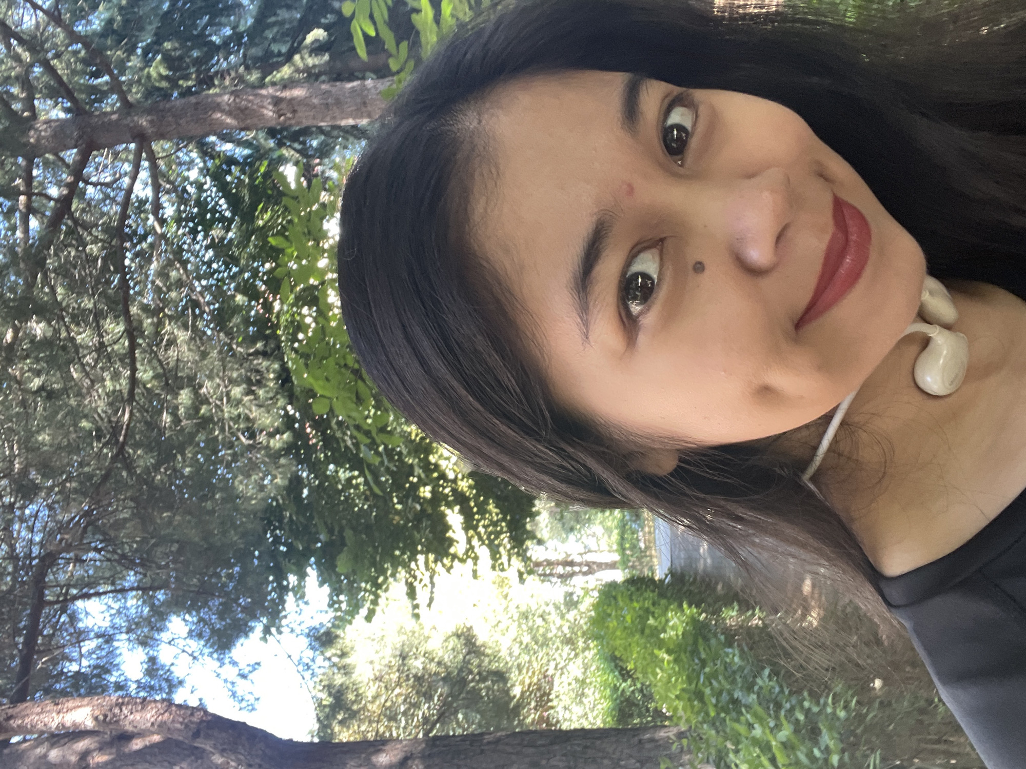 A woman with long dark hair and fair skin smiles at the camera in an outdoor setting. She has dark eyes, red lipstick, and a beauty mark on her left cheek. She is wearing a black shirt and white earbuds around her neck. The background shows trees, greenery, and a path, suggesting a park or garden. The lighting indicates it is daytime. The image was taken in the city of Başakşehir, Turkey.