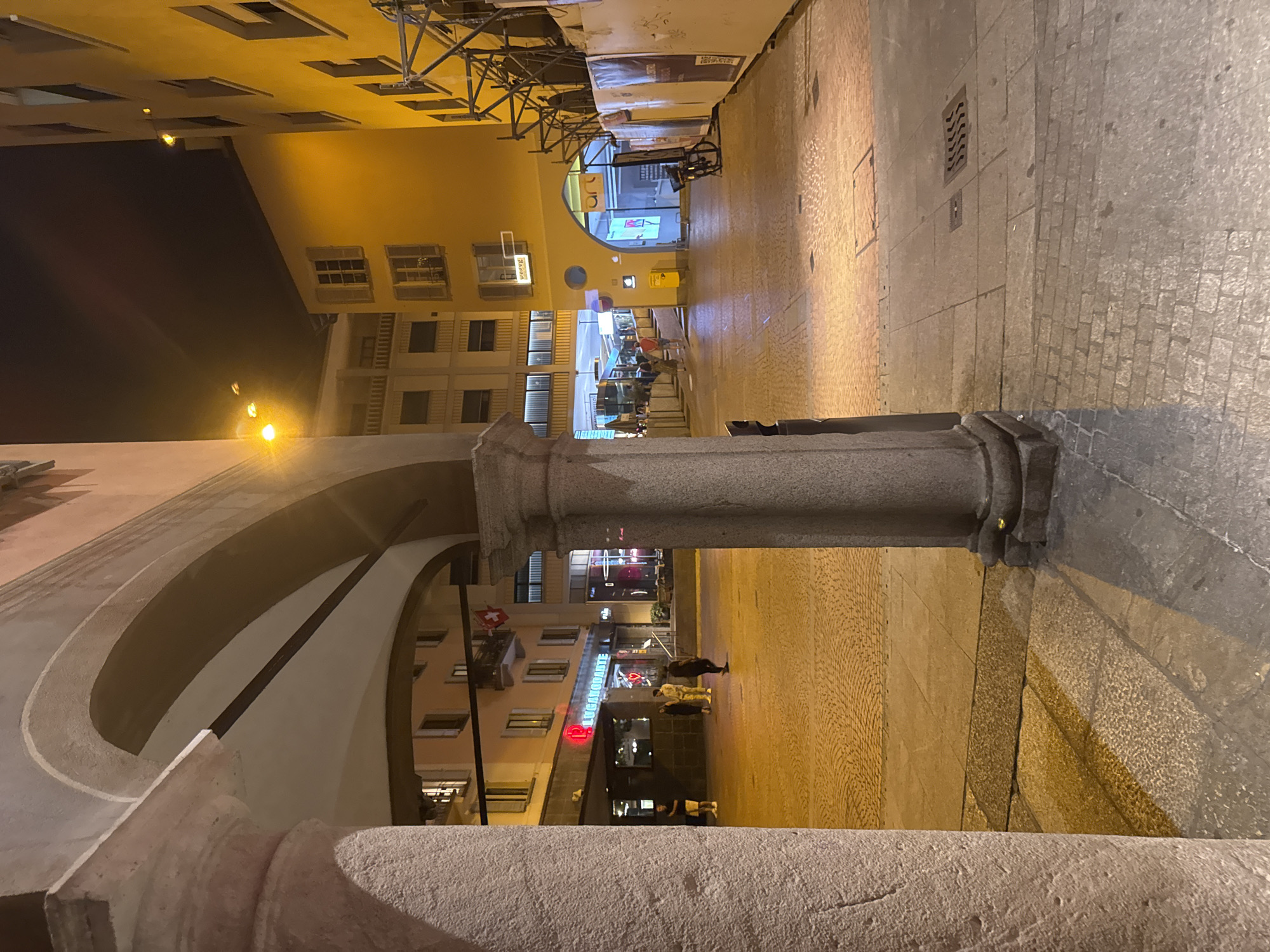 The image shows a night view of a pedestrian area in Lugano, Switzerland, likely near Piazza della Riforma. In the foreground, a large stone column and archway frame the scene. The ground is paved with cobblestones and stone slabs, illuminated by streetlights and the warm glow from building interiors. Several people are visible walking in the distance. To the left, a building with a red neon sign displaying "PUCCIANTE" and a heart symbol suggests a restaurant or establishment. Further down the street, another building has a sign that reads "ART" above an arched entrance. Scaffolding is present on the right side of the image, indicating ongoing construction or renovation. The overall atmosphere is calm and European, typical of a city square at night.