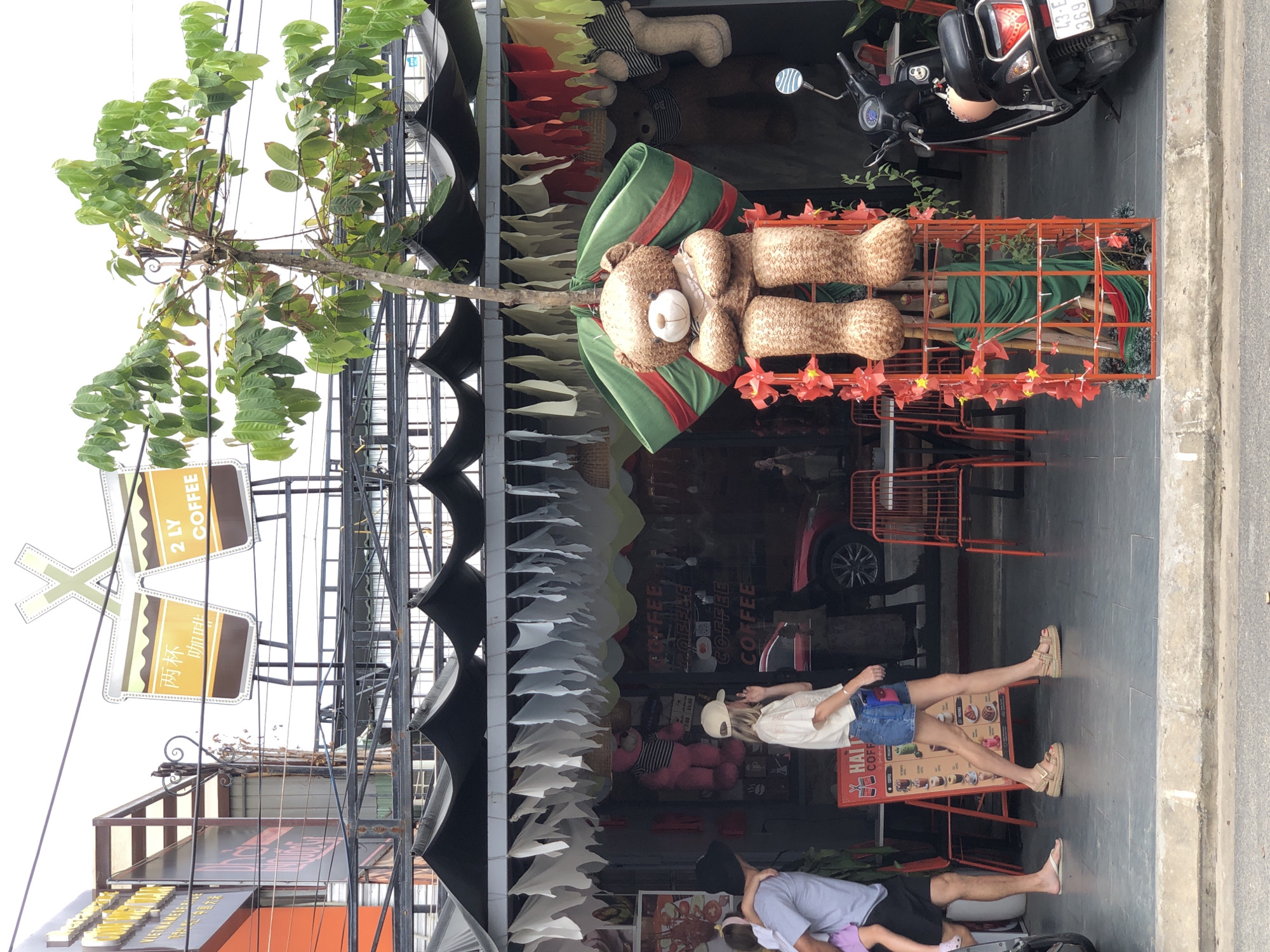 This is a medium-shot, eye-level, outdoor photograph taken in the city of Đà Nẵng, Vietnam, on a street lined with businesses and decorated with various items. In the foreground, a large teddy bear statue is prominently displayed. The bear is seated and wearing a green and red sash draped around its shoulders. It is enclosed in a decorative orange metal cage adorned with red paper pinwheels and green fabric. To the left of the teddy bear, a woman in a white t-shirt, denim shorts, and sandals is walking. She is wearing a beige baseball cap and has her right hand near her face, possibly touching her ear or hair. To her left, a man wearing a dark t-shirt and black shorts is holding a young child from behind. The child, wearing a purple garment, is facing away from the camera. Behind them, a red sign with text and images of drinks is visible. Further back, there is a storefront with a black awning supported by metal poles, decorated with white and red hanging fabric strips. The storefront displays multiple pink teddy bears. A red car is parked inside the storefront. On the right side of the image, a black scooter with a license plate that reads "43-E1 369.70" is parked. The environment appears to be a pedestrian walkway in front of shops on a street. The sky is overcast and appears white. The signage above the awning features two stylized coffee cups and text in Vietnamese and English that reads "2 LY COFFEE."