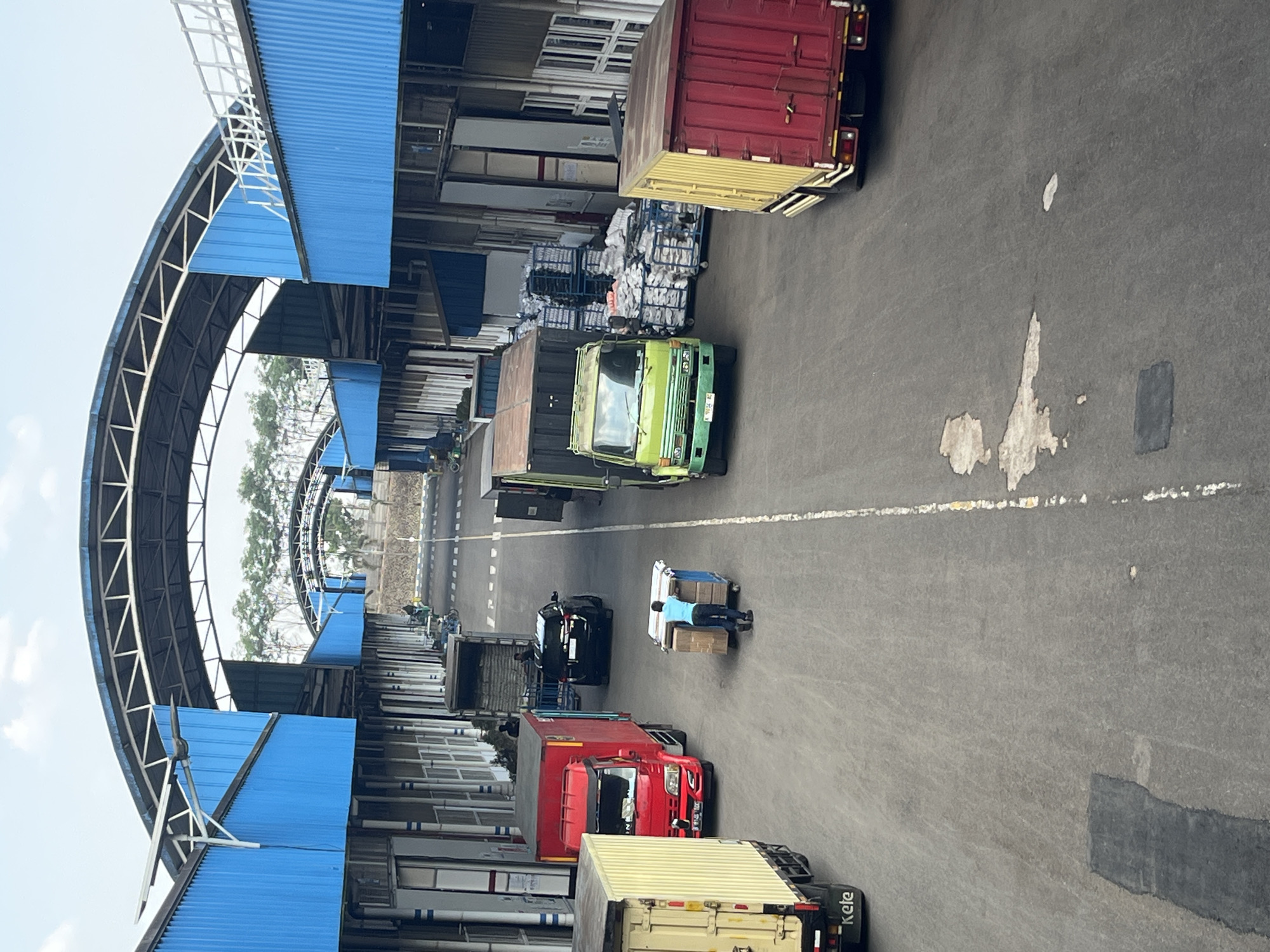 The image depicts an outdoor loading dock or distribution area. A person in a blue shirt is pushing a cart loaded with boxes. Several trucks are present: a red truck, a green truck, a yellow truck, and a red and yellow truck. The scene is framed by industrial buildings with blue roofs and siding, suggesting a commercial or logistics facility. The ground is asphalt, marked with white lines. Trees are visible in the background, and the sky is overcast. The location is identified as Pelang, Indonesia, which is a city in East Java, Indonesia, known for its agricultural and industrial activities.
