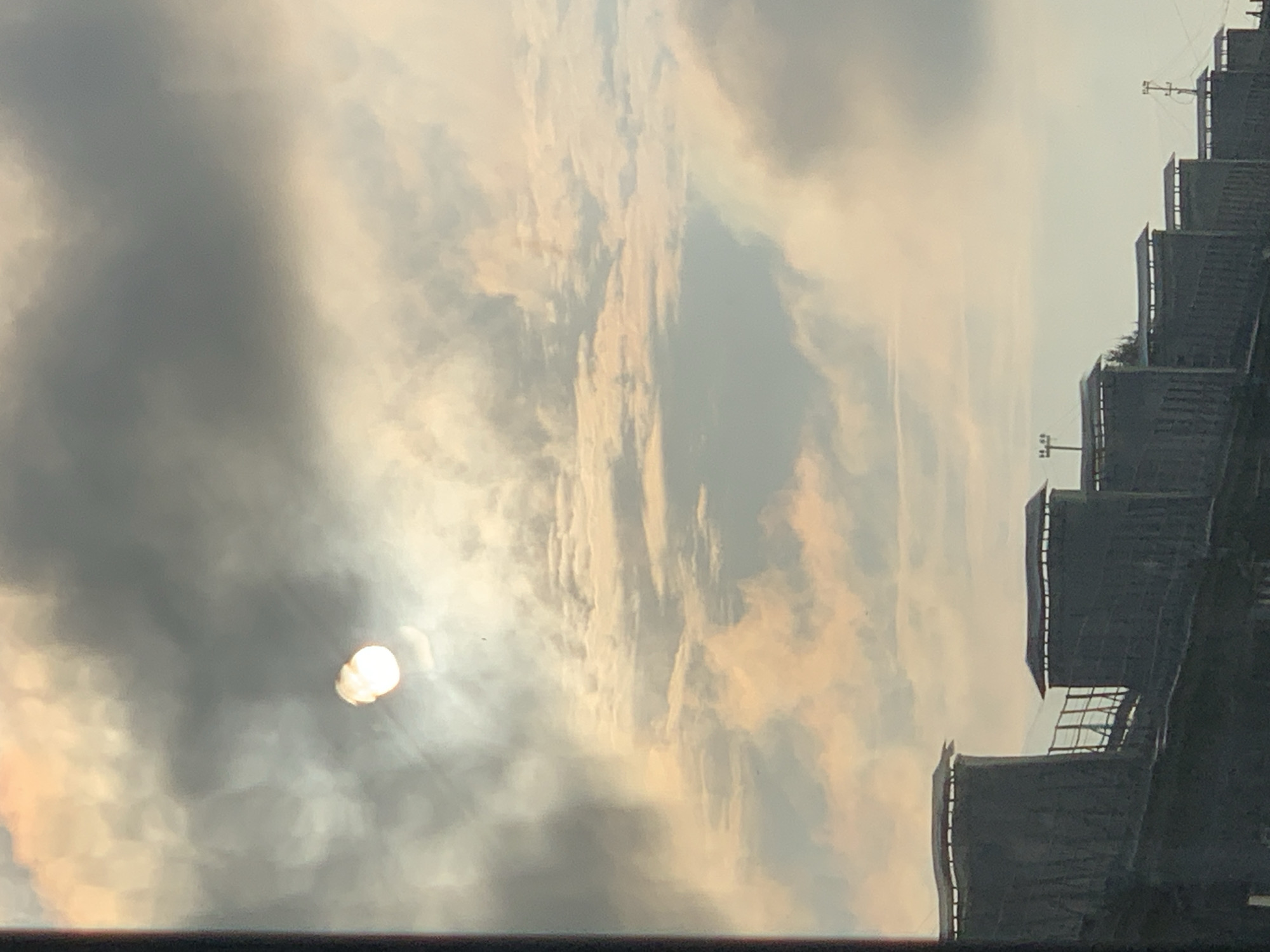 The image captures a cloudy sky with the sun partially visible through the clouds, creating a diffused glow. Below the sky, a row of identical, dark-colored structures with peaked roofs are lined up. These structures appear to be part of a building complex or industrial facility. The overall scene is overcast with a hint of warm light from the sun, suggesting it might be sunrise or sunset. The location is identified as Prokopyevsk, Russia.
