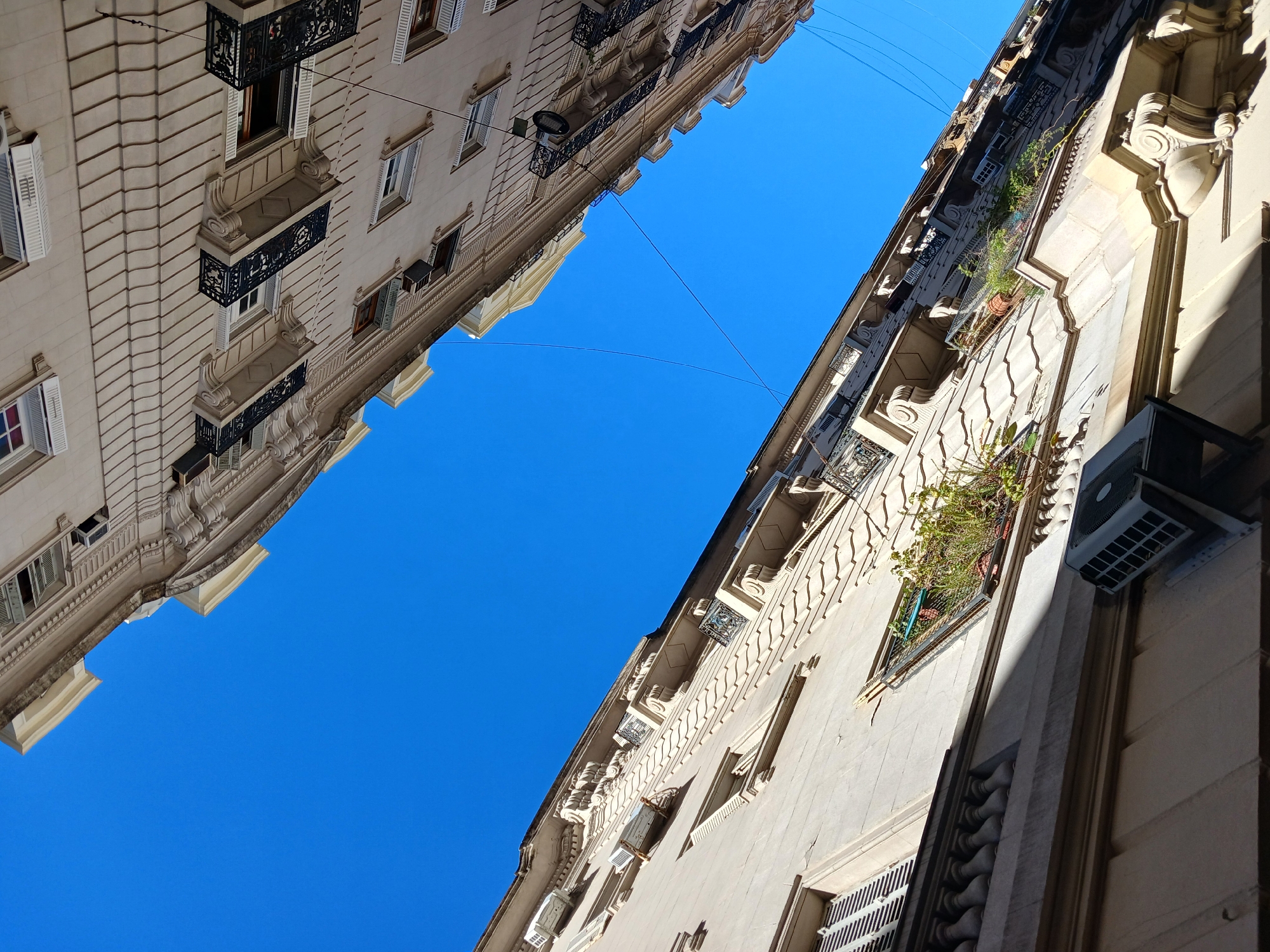 The image captures a view of two buildings in Buenos Aires, Argentina, angled upwards against a clear blue sky. The buildings are traditional in style, featuring light-colored facades, multiple windows, decorative balconies, and detailed molding. Air conditioning units are affixed to the exterior of one building. Overhead wires are visible against the sky, connecting the two structures. No people are visible.