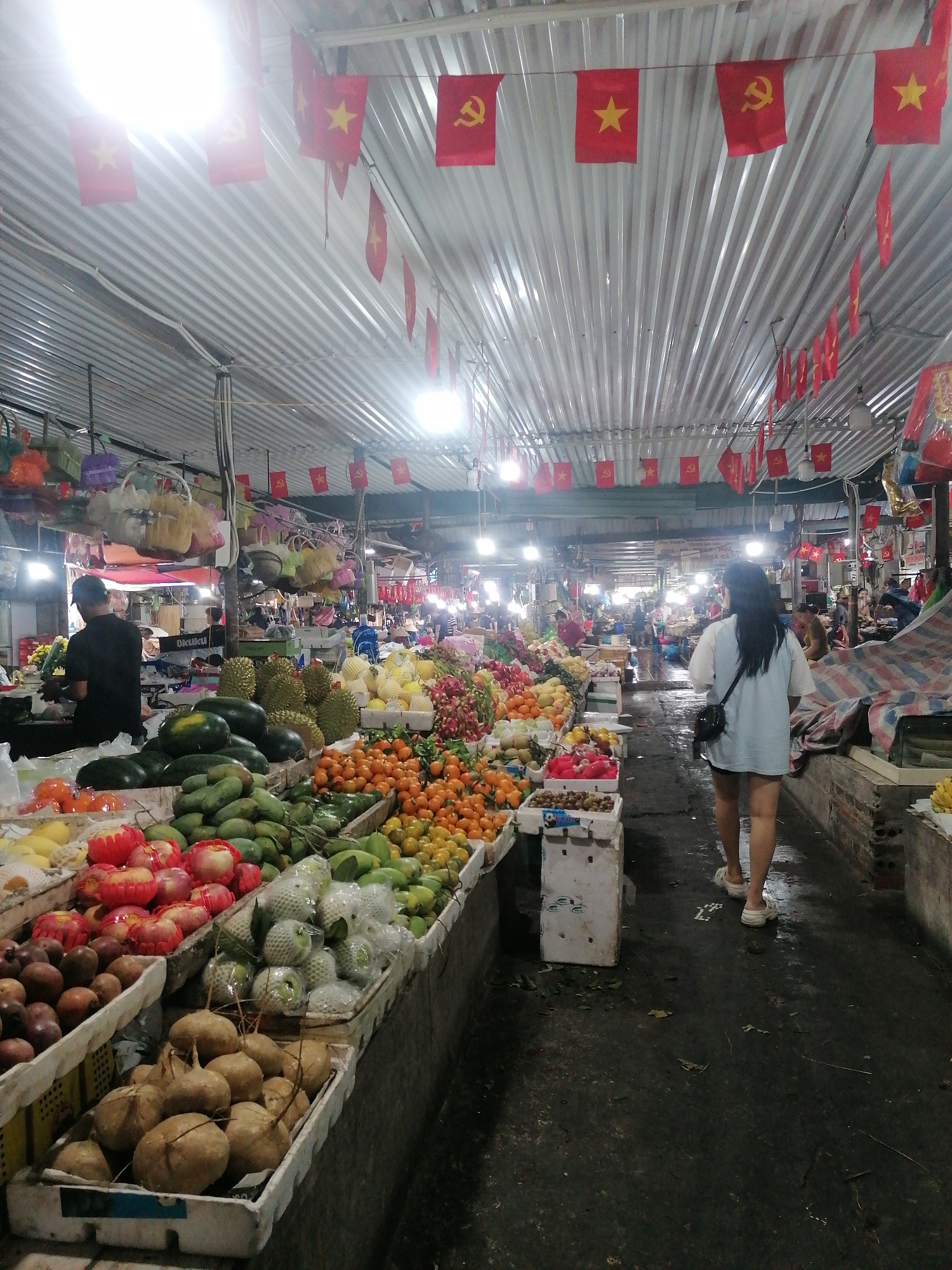 This image captures a vibrant indoor market scene, likely a fruit market, in Bai Chay Ward, Vietnam. The setting is characterized by a long, covered aisle with stalls lining both sides, all beneath a corrugated metal roof. Numerous small red flags, featuring both a yellow five-pointed star (representing the national flag of Vietnam) and a yellow hammer and sickle (symbolizing the Communist Party of Vietnam), are strung across the ceiling, serving as prominent location cues.
The market stalls are laden with a diverse and colorful array of fresh produce. In the foreground and mid-ground, there are large displays of fruits such as watermelons, spiky durians, green mangoes, tangerines, and various other items including what appear to be jicama, grapes, and guavas (some individually wrapped). Decorative red items, possibly artificial apples or lanterns, also adorn one of the fruit displays. Overhead, colorful woven baskets hang, adding to the market's bustling atmosphere.
Several people are visible within the market. On the left side, a person wearing a black shirt and cap stands behind a stall, possibly a vendor attending to customers. In the right mid-ground, a woman with long dark hair, dressed in a light-colored loose top and dark shorts, walks away from the viewer down the central aisle, carrying a black shoulder bag. Other individuals can be seen in the background, further along the market, suggesting a busy environment.
The floor of the market appears dark, wet, and somewhat uneven, typical of a fresh produce market. Bright artificial lights, likely fluorescent or LED, are suspended from the ceiling, illuminating the stalls and their wares. The overall impression is one of a lively, functional marketplace. Visible text is minimal; "OKUKU" is faintly discernible on a stand in the background, and some illegible text appears on white boxes.
