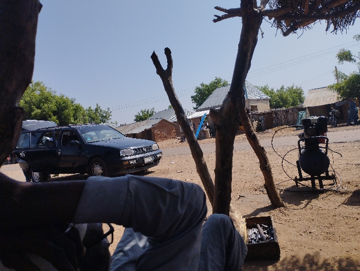 The image was taken in Tongo, Nigeria (10.664, 11.364). It shows a partially obscured view of a person sitting under a large tree in a dusty, dirt-road setting. The person's legs are visible, clad in dark pants and a dark shirt. The tree's branches stretch out, partially obscuring the view of the surroundings. A black car is parked in the foreground on the road. The car is a Volkswagen Golf, and it's facing away from the camera. A metal object, potentially a compressor, is sitting on the ground to the right of the car. In the background, there are a few structures and buildings that appear to be in a residential area. There are trees on the left side of the image and some palm trees in the background. The image appears to have been taken in daylight on a clear day.