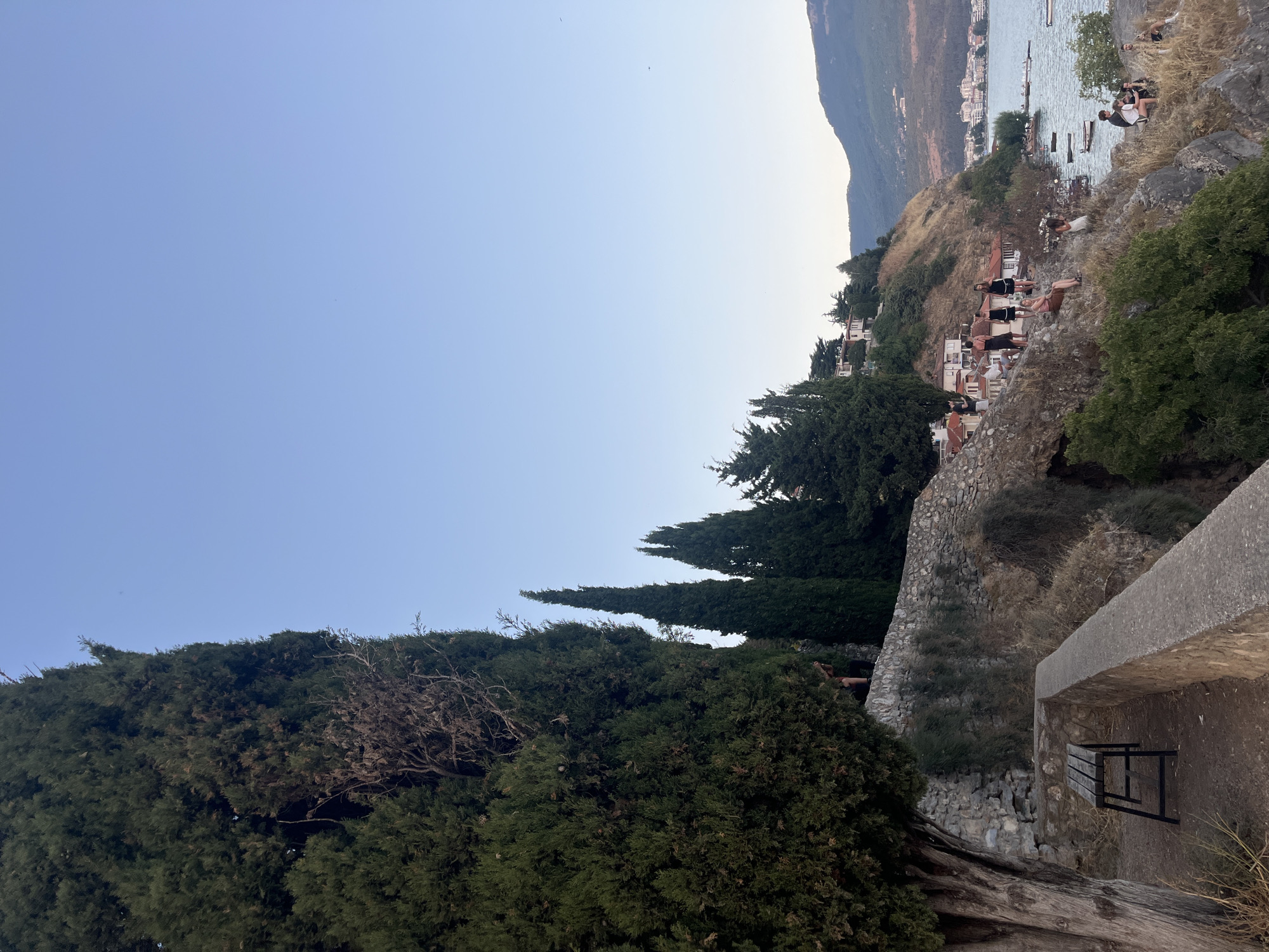 The image depicts an outdoor scene with a focus on a cliffside overlooking a body of water. A large evergreen tree dominates the left side of the frame, with a rustic stone wall and a wooden bench in the foreground. Several individuals are scattered along the rocky incline, some sitting, others standing. They appear to be engaged in conversation or simply observing the view.
Further down the cliff, a cluster of buildings with red-tiled roofs is visible, suggesting a small settlement. The water in the foreground is calm, with a few small boats moored near the shore. In the distance, a larger town or city is discernible along the coastline, nestled against a backdrop of rolling hills. The sky is a clear, pale blue, indicative of daylight. The overall impression is one of a scenic overlook offering a panoramic view of a coastal landscape.