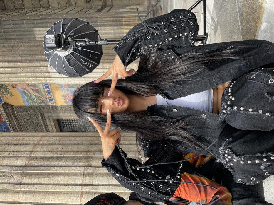 An East Asian-appearing female is positioned centrally, crouching or sitting outdoors in front of the Panthéon in Paris, France. She faces the viewer, winking her left eye, pursing her lips, and making "peace" signs with both hands to the sides of her face. Her long, straight dark hair with bangs is visible. She has a bridge piercing and long, multicolored decorated fingernails (light blue/green with glitter on the left index, pink on left middle; pink on right index, light blue/green on right middle). She wears a black denim jacket and matching pants, both extensively embellished with silver studs and black lacing through eyelets, giving a corset-like appearance on the sleeves and pant seams. A white crop top is visible beneath the jacket. To her left, part of another individual wearing a dark cap and an orange and black patterned garment is visible. To her right, a black Aputure Light Dome parabolic softbox with a strobe head is mounted on a stand, positioned for a photoshoot. The background consists of the light-colored stone facade of the Panthéon, featuring large fluted columns. Between the columns, an ornate section above a dark recessed doorway displays a colorful mural depicting a partially nude male figure in a landscape with the legible text "CHUTE D" below it, and further down, additional mural elements with winged figures.