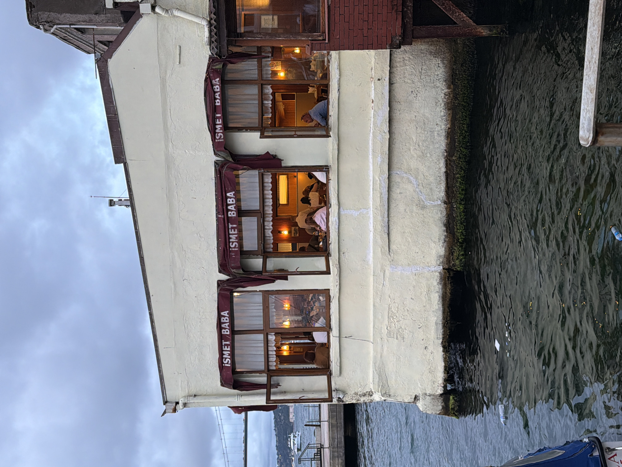 The image captures the exterior of a building situated on the waterfront in Üsküdar, Turkey. The building, painted a light cream color, features multiple windows that look into an interior space, possibly a restaurant or cafe, indicated by the presence of people seated at tables and ambient lighting. The windows are adorned with burgundy awnings that bear the text "İSMET BABA".
To the left of the building, the Bosphorus strait is visible, with a boat sailing in the distance and a suspension bridge, likely the Bosphorus Bridge, spanning across the water. The water itself appears somewhat choppy and carries a small amount of debris, including plastic bottles and wrappers. The building's foundation is in contact with the water, and marine growth is visible along the waterline.
To the right, a section of the building constructed with red brick is visible, with supporting wooden structures indicating a pier or an elevated platform. The sky above is overcast, suggesting a cloudy day. The overall scene depicts a slice of urban waterfront life in Üsküdar.