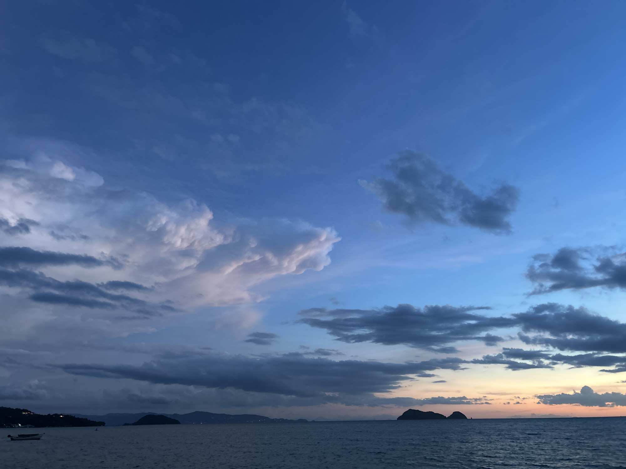 The image depicts a scenic coastal view at dusk in Ko Pha-ngan District, Thailand. The sky is a gradient of deep blue near the top, transitioning to lighter blues and hints of orange and yellow near the horizon, indicating the setting sun. Scattered clouds are visible, some dark and ominous, while others are illuminated by the fading light, appearing soft and white.
The sea occupies the lower half of the frame, appearing calm with gentle ripples on its surface. In the distance, a silhouetted coastline is visible, with rolling hills and mountains. A small, dark, irregularly shaped island or rock formation is situated in the middle-right of the sea, with two smaller, similar formations slightly to its left.
On the far left of the image, a small, white boat with a dark hull is anchored on the water, close to the shore. The coastline on the left appears more developed, with lights suggesting a settlement or buildings. The overall atmosphere is serene and tranquil, characteristic of a twilight scene by the sea. There are no people or animals clearly visible in the image.