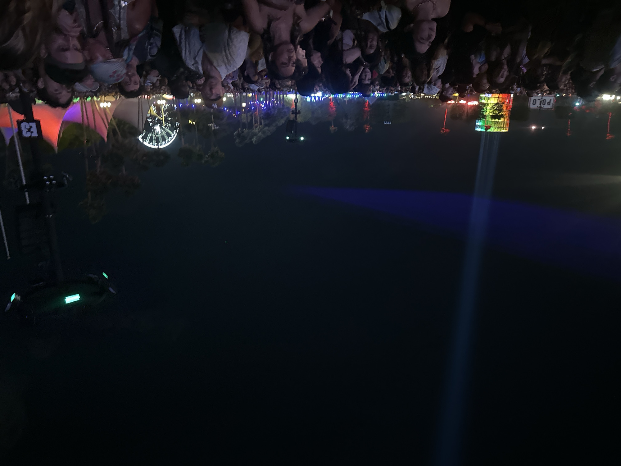 The image shows a nighttime view of a crowd of people at a festival in Unknown, Unknown. The crowd is densely packed and appears to be facing a stage or focal point, which is indicated by a large beam of light projecting upwards. Various structures, including a Ferris wheel, colorful tents, and a sign that reads "POLO," are visible in the background. The lighting suggests it is a night event, with various colored lights illuminating the scene.