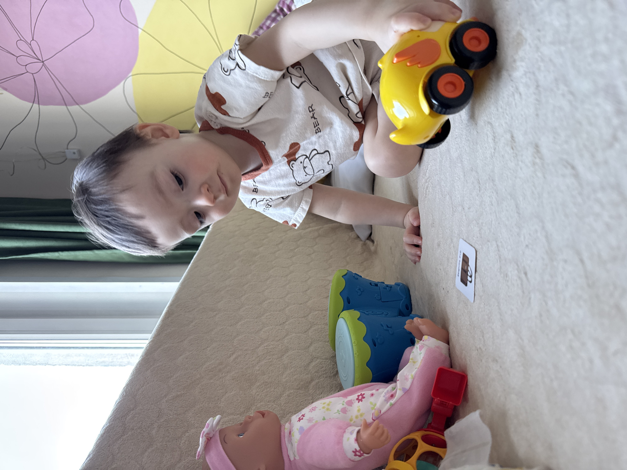 A young child is positioned on a light-beige surface, possibly a bed or sofa. The child, wearing a white shirt patterned with brown bears, holds a yellow toy car with orange wings and black wheels. A doll dressed in pink lies near a blue and green toy. Adjacent to these toys, a card with an image of a brown bag is visible. Above the child, sections of a yellow and pink object are visible. Part of a window is to the left of the child. The location is not discernible.