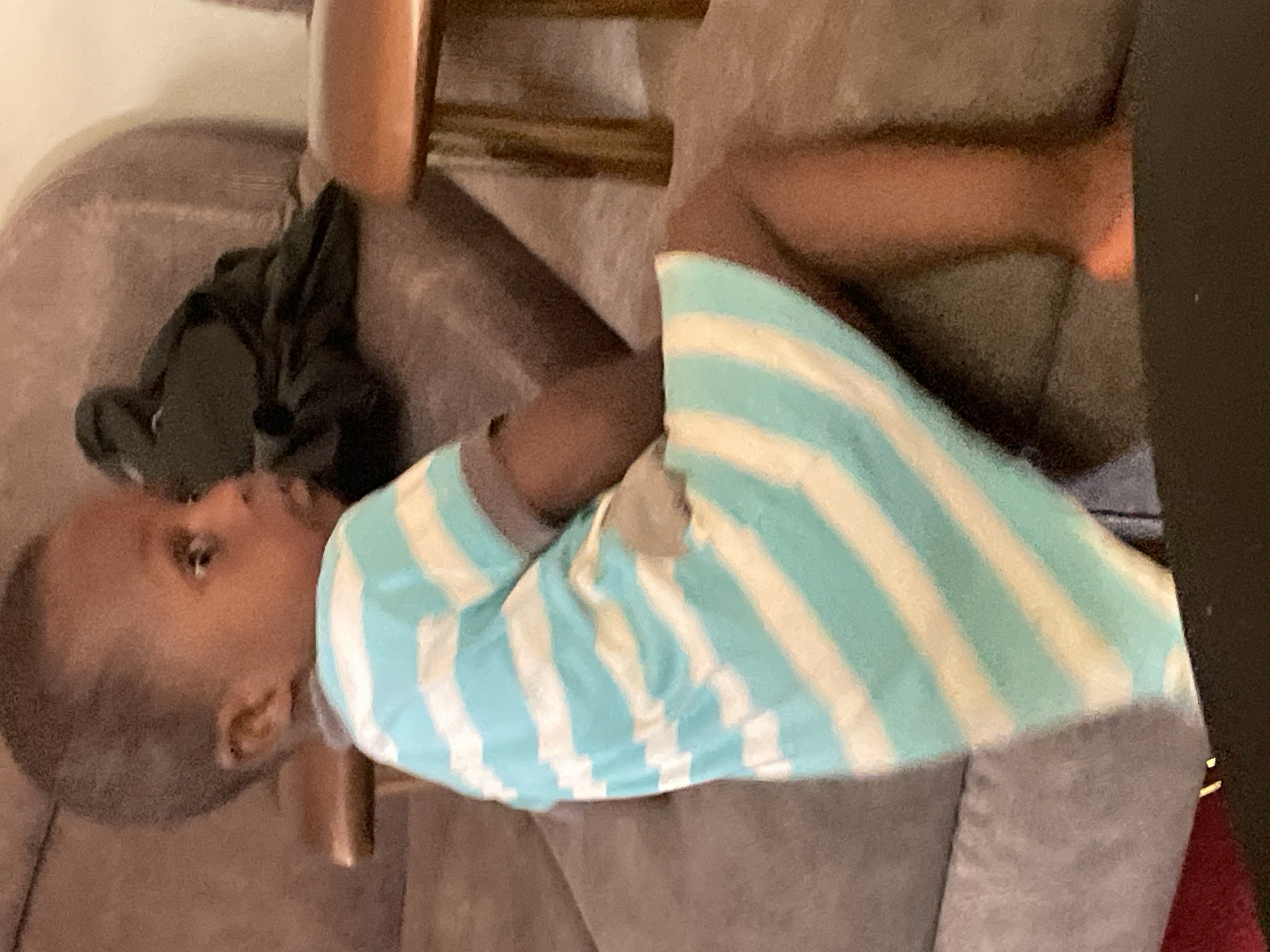 A child with dark skin and short, dark hair is lying on a brown sofa. The child is wearing a blue and white striped shirt. The child is looking to the left. The location is Jos, Nigeria. The child's arm is resting on the sofa. There is a black garment next to the child. A portion of a chair is visible in the background.