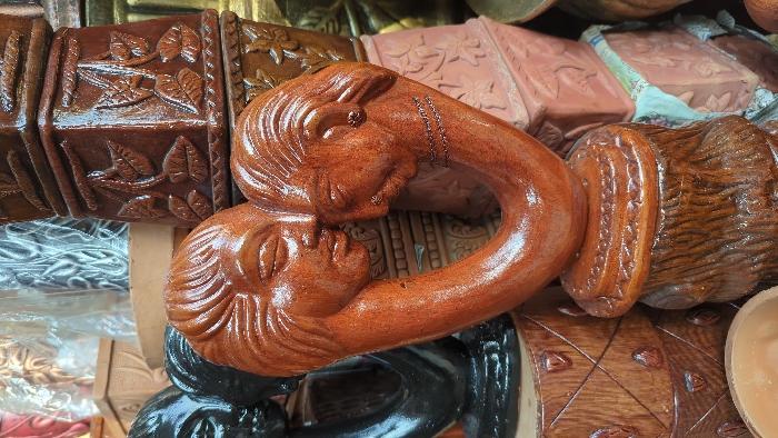 The image presents a close-up view of a retail display containing various terracotta and clay items. Dominating the center is a reddish-brown, glazed terracotta sculpture featuring two stylized human faces in profile, positioned to face each other with their foreheads and noses in near contact. This central sculpture forms a U-shape, resting on a textured, cylindrical base with a decorated rim. Both sculpted faces exhibit closed eyes and stylized hair; the right face additionally shows an ear adorned with a small, round stud and etched three-strand necklace details on the neck.
The background and surrounding areas are densely packed with other pottery. To the immediate left of the central sculpture, a dark, possibly black-glazed terracotta sculpture of a human head is partially visible. Behind and to the right, multiple brown-glazed square or rectangular pots are stacked, each featuring embossed leaf patterns. Further back and to the upper left, lighter-colored pots with various patterns, some appearing to be wrapped in clear plastic, are visible. Fragments of newspaper or light-colored fabric are interspersed among the stacked items, likely used for cushioning or display. The overall environment suggests an outdoor market stall or pottery shop. The specific location within Dhaka, Bangladesh, cannot be determined from the visual information provided.