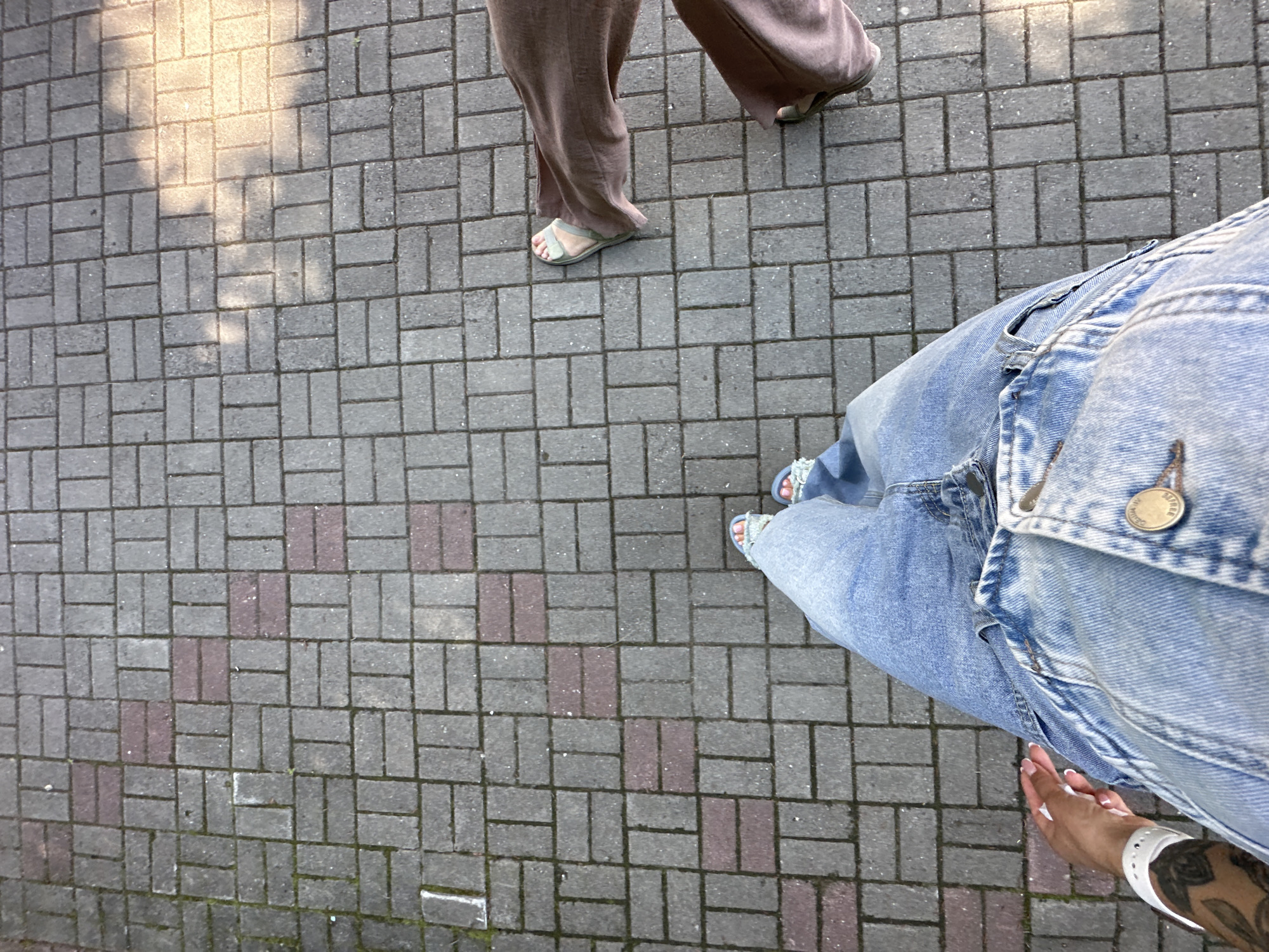 The image shows a top-down view of two individuals standing on a gray brick pathway. The person in the foreground is wearing a denim jacket and light blue jeans, with their left hand visible on their left hip. They have a white wristband on their left wrist and visible tattoos on their arm. The second person is partially visible on the right, wearing brown wide-leg pants and sandals. The pathway has some red bricks interspersed with the gray ones and is partially covered in sunlight, casting shadows. The environment suggests an outdoor urban setting, possibly a park or a pedestrian walkway in Duvankul', Russia, given the context provided.